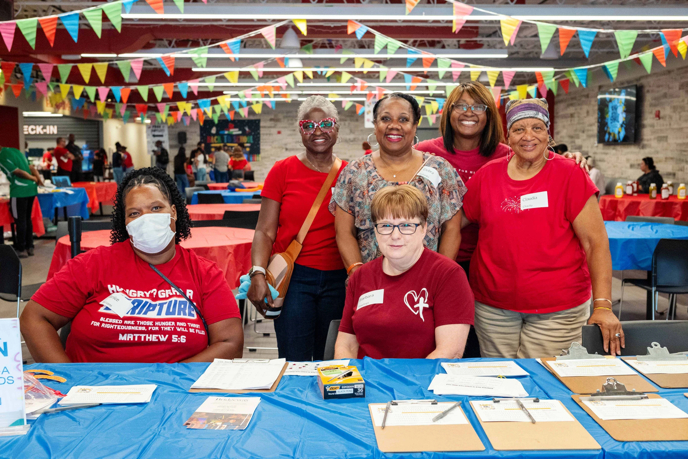 A group of seven smiling women volunteers posing for a picture at a registration table in a brightly decorated hall with colorful banners.