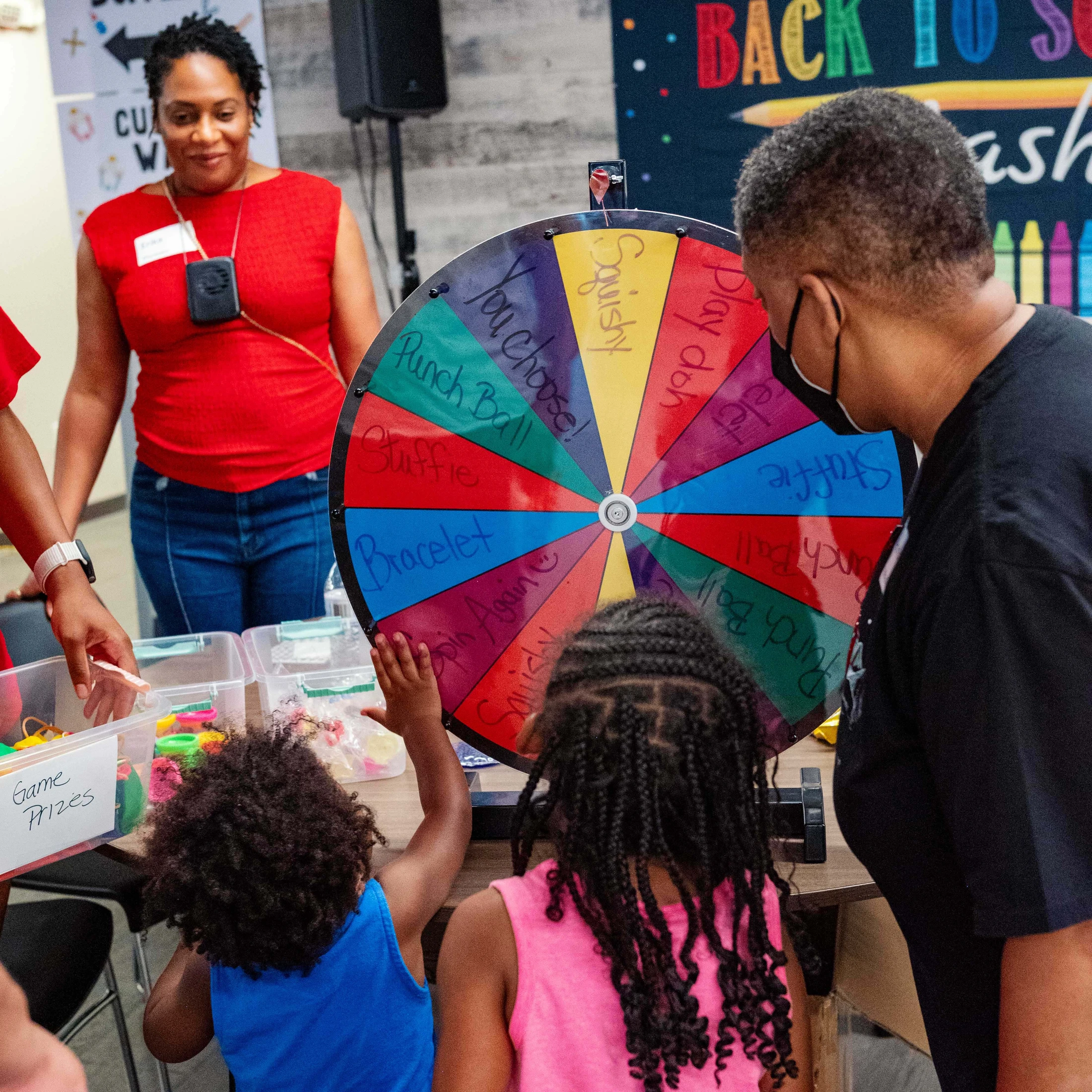 Two young children reach out to spin a colorful prize wheel at a back-to-school event while two adults watch them.