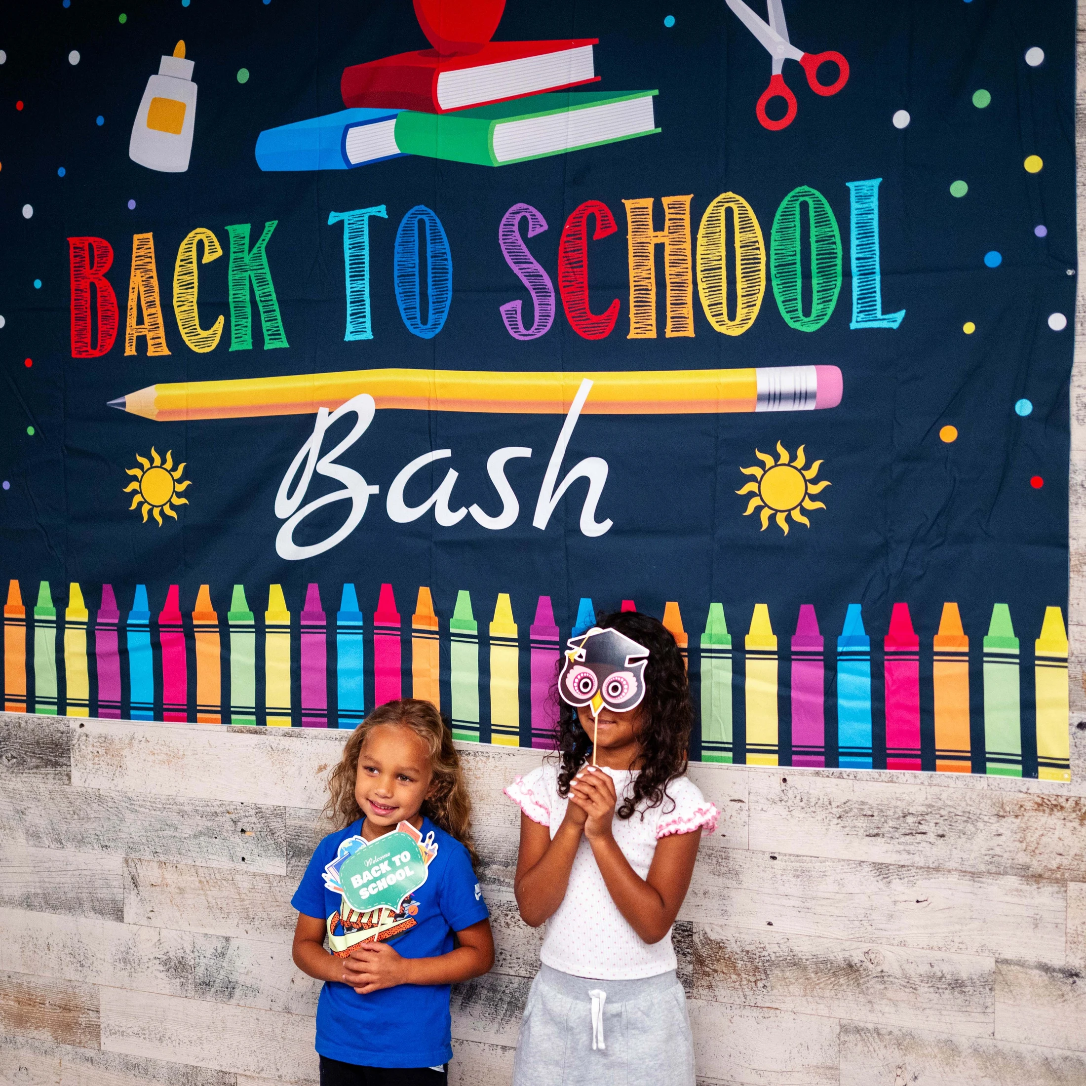 Two young girls holding photo props stand in front of a colorful 'Back to School Bash' banner with illustrations of school supplies.