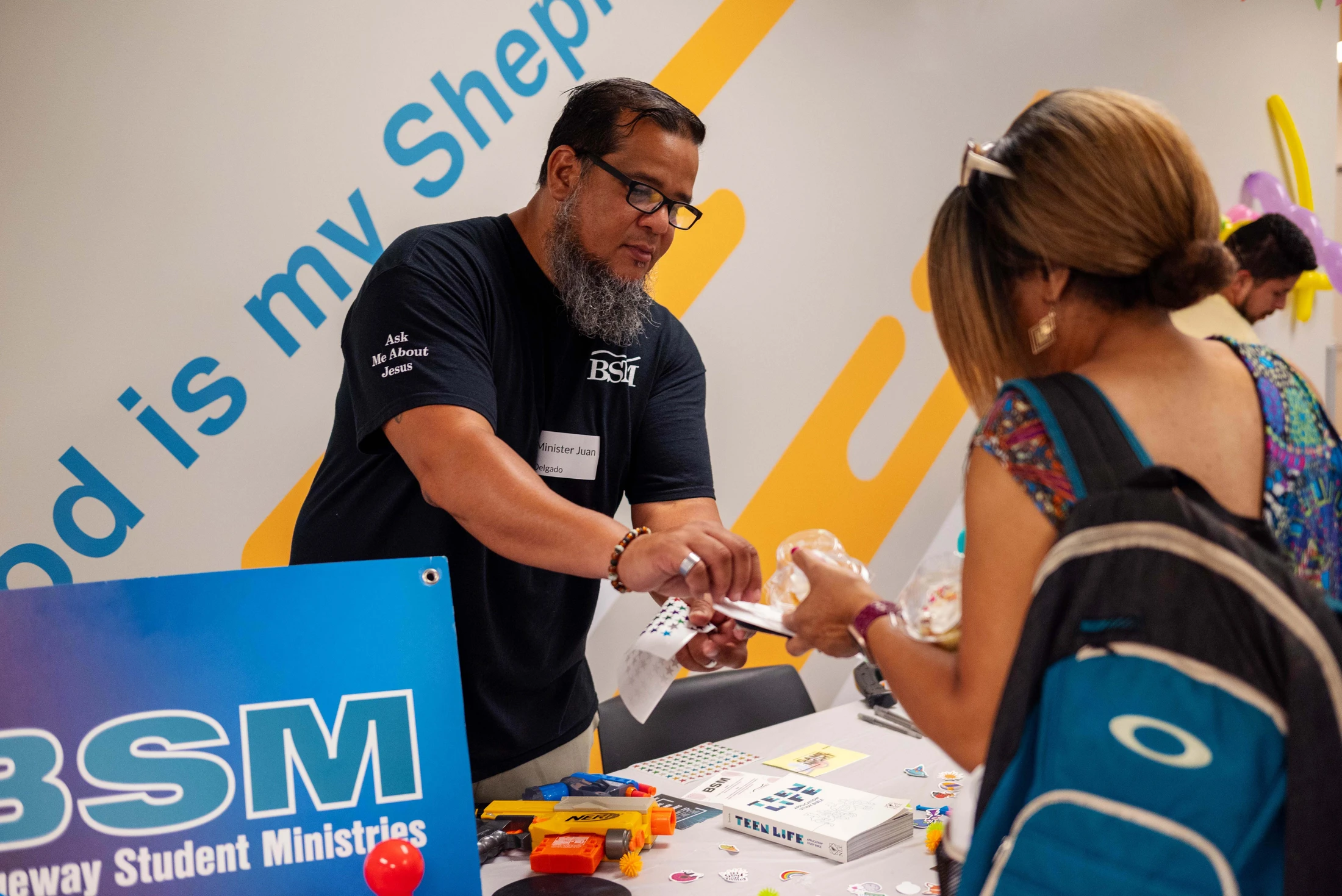 A minister with a beard and glasses hands an item to a woman across a table at a Blueway Student Ministries event.
