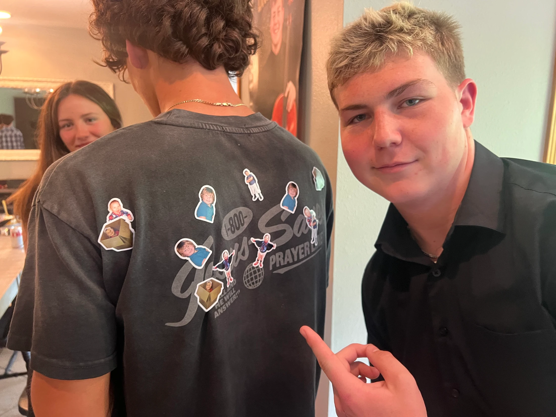 A young man points and smiles at the back of a friend's shirt, which is covered in custom stickers of his embarrassing childhood photos.