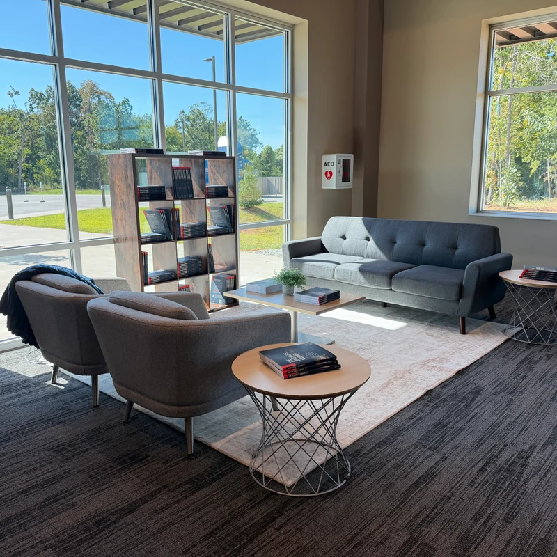 A sunlit waiting area with modern grey furniture, a large window, and a bookshelf. Sunlight casts shadows across the dark grey carpet and cream-colored rug.