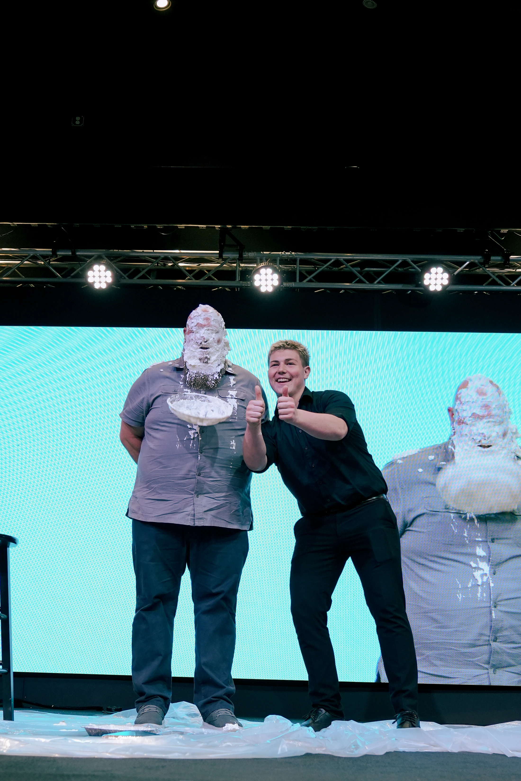A smiling young man gives two thumbs up, standing between two men with their faces covered in cream from a pie during a fun event on a stage.