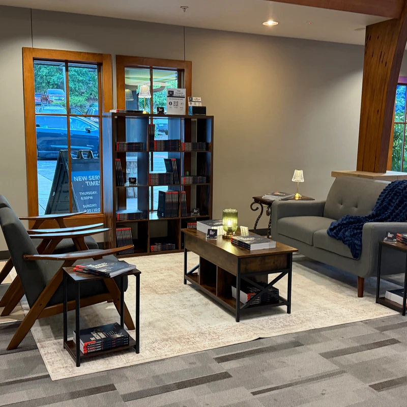 A modern and cozy seating area in a lobby with a gray sofa, an armchair, a coffee table, and a bookshelf filled with books called 'The Disciple's Journey'.