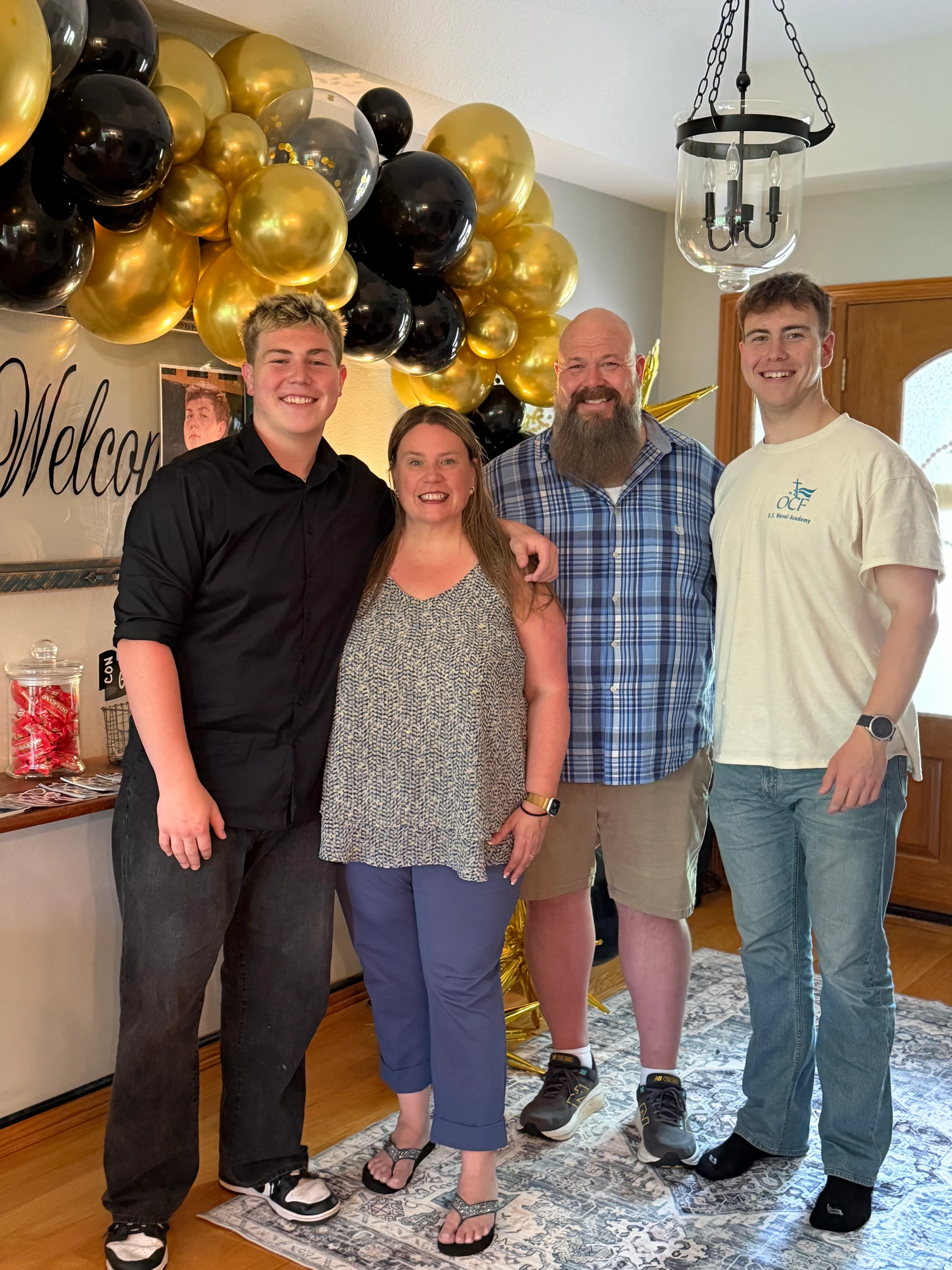 A family of four, parents and two sons, smile for a photo in their home. In the background, there is a large arrangement of black and gold balloons.