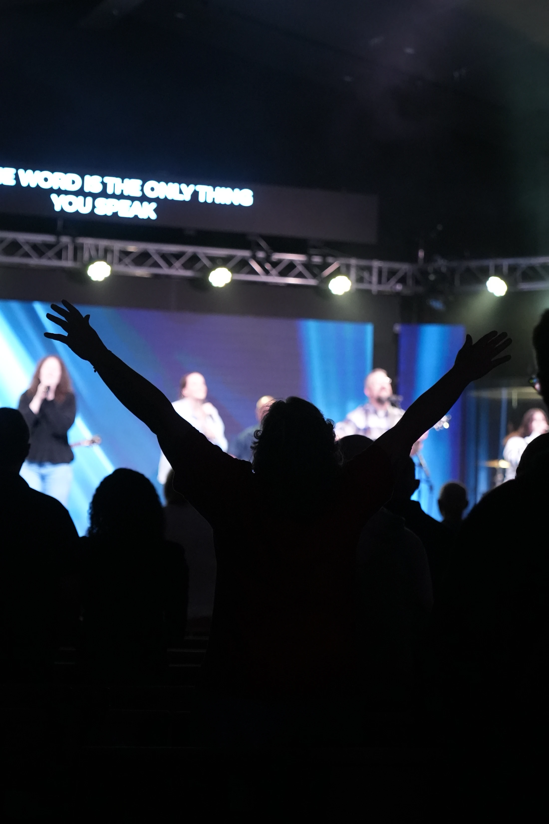 A silhouette of a person with arms raised in worship, facing a brightly lit stage where a band is performing. A screen above displays song lyrics.