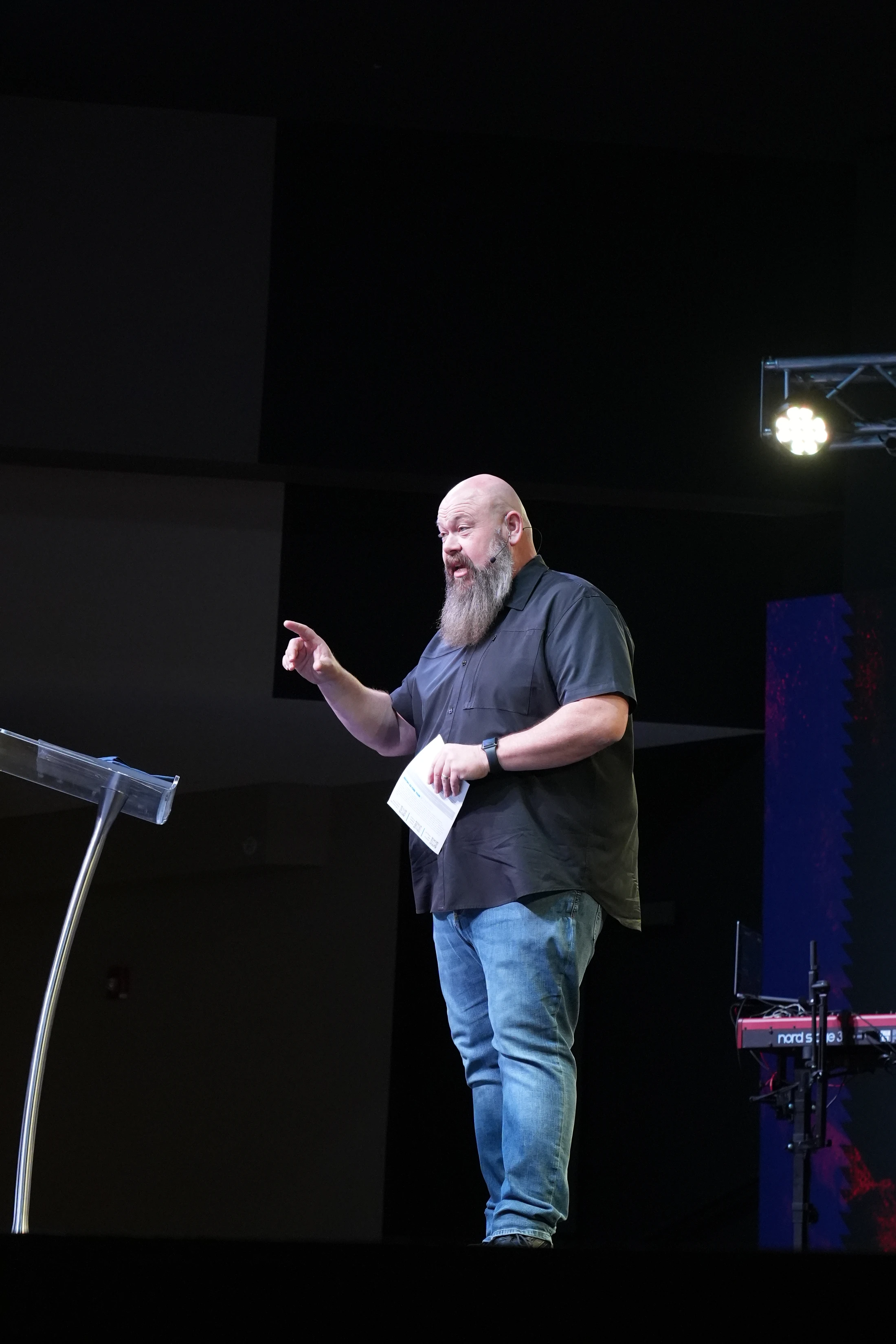 A bald man with a long grey beard stands on a dark stage, wearing a black shirt and jeans. He is speaking and pointing while holding a paper.