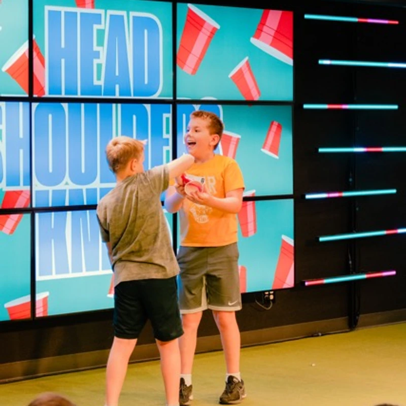 Two young boys compete on a brightly lit stage to grab a red cup, with an audience watching and a large screen behind them displaying the name of the game.