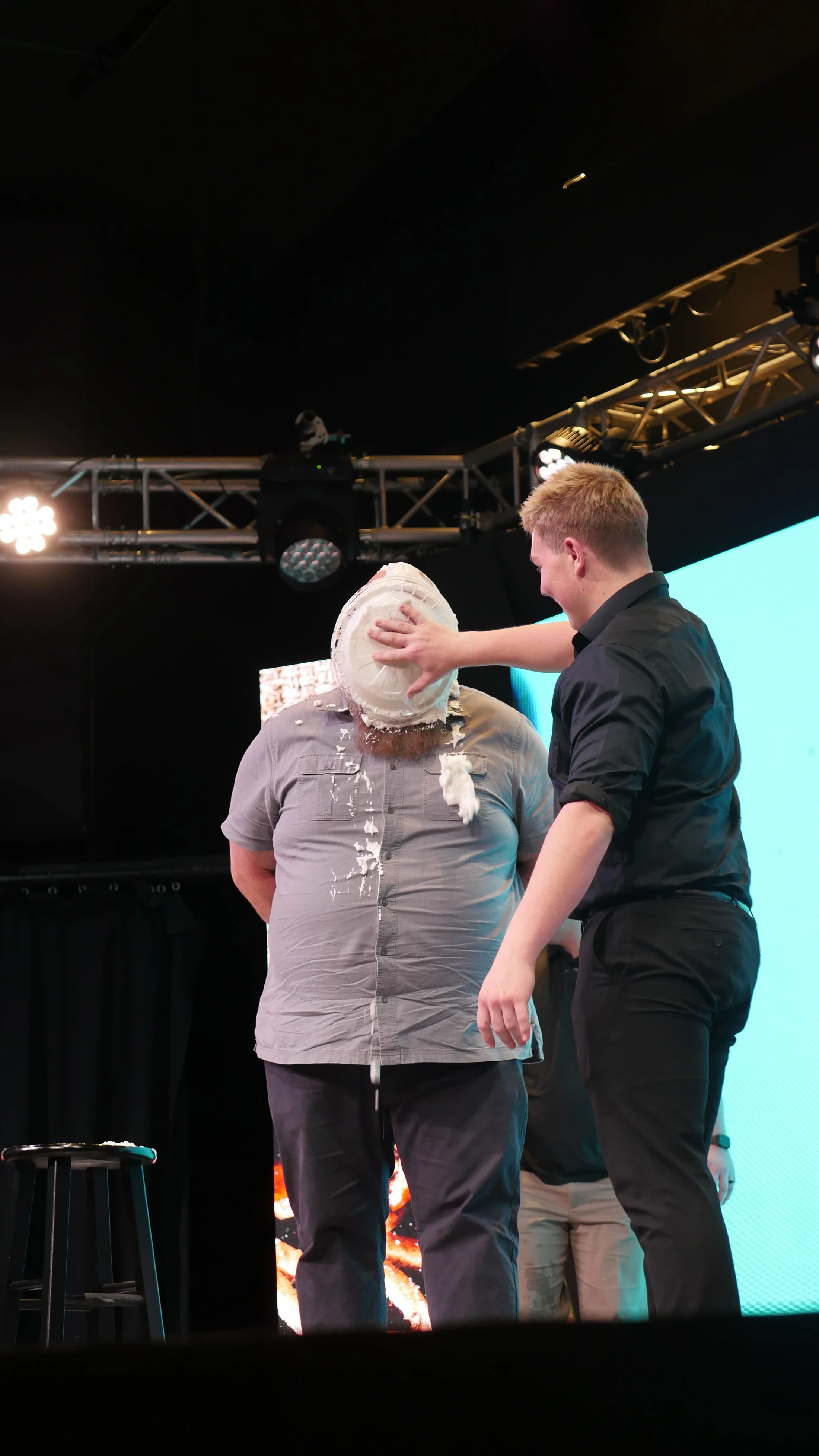 A man in a black shirt smiles as he pushes a pie tin full of whipped cream into another man's face on a stage, splattering cream onto his grey shirt.