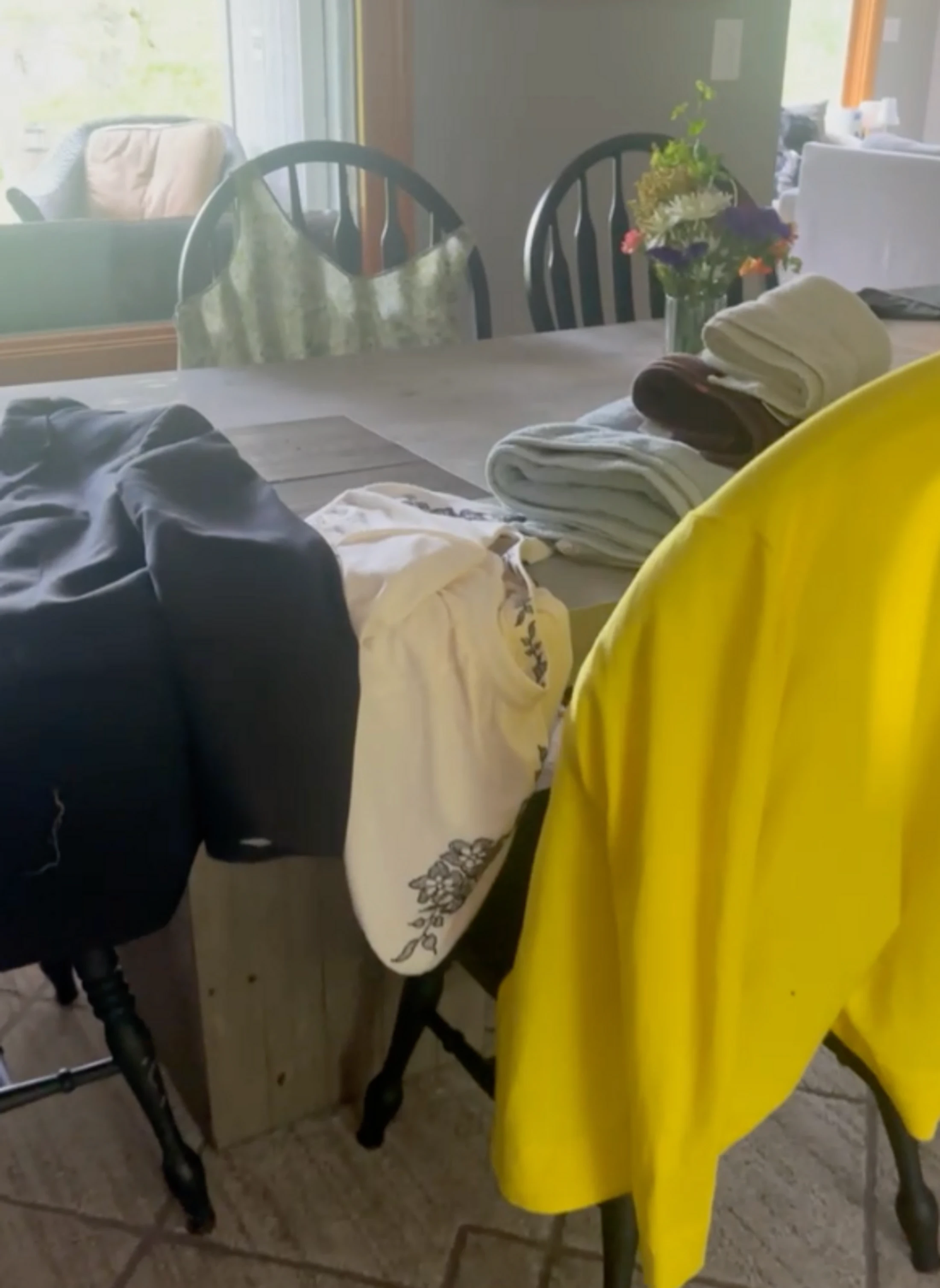 A cluttered dining room with various items of clothing draped over black chairs and folded towels stacked on the wooden table next to a vase of flowers.