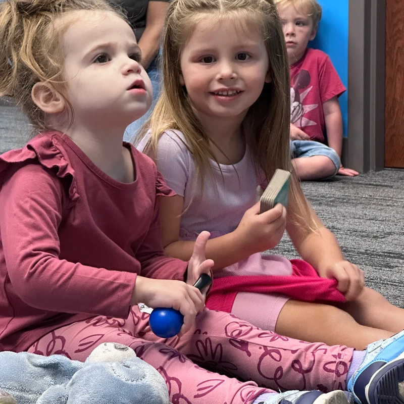 Two young girls sit on a gray carpet. One looks up attentively while the other smiles at the camera. A little boy sits in the background.