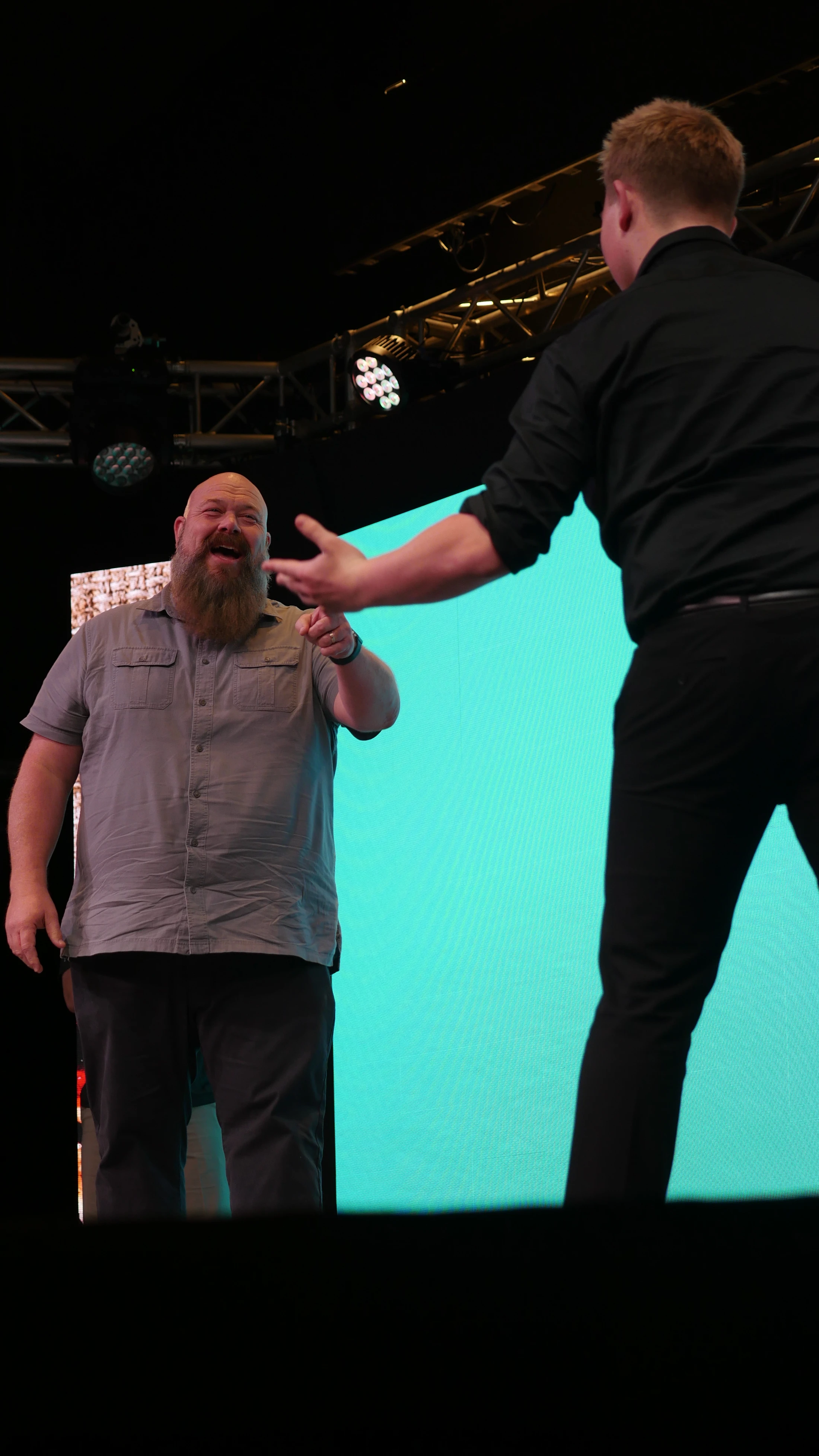 A bald man with a large beard smiles and points at another man on a stage. They stand in front of a large, bright teal screen with lighting rigs above.