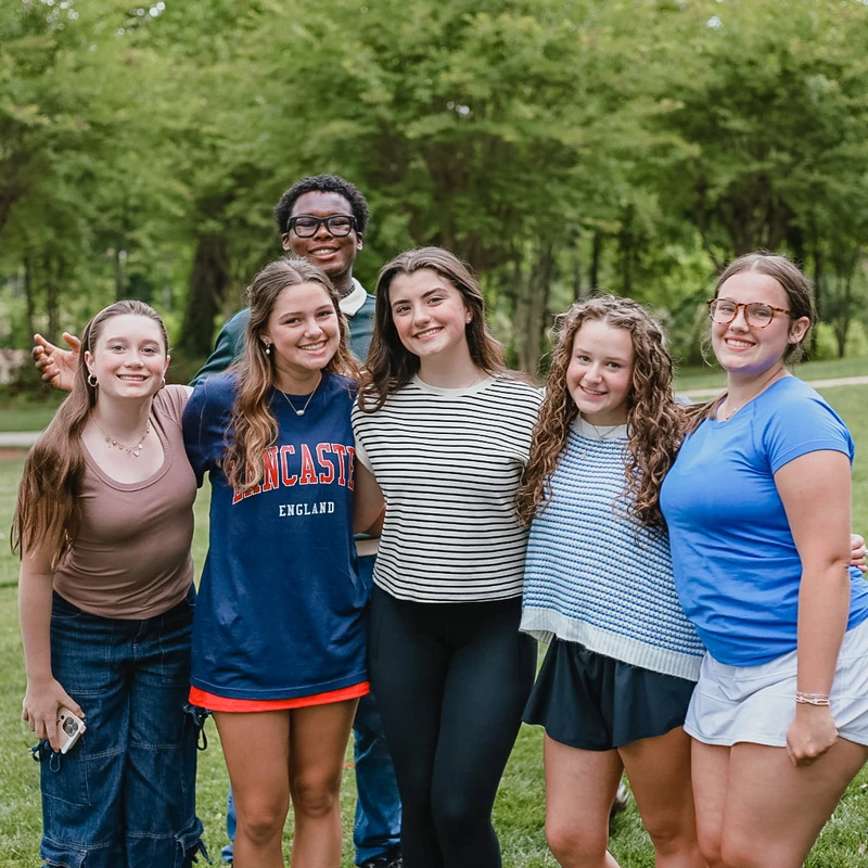 A diverse group of six teenagers, five girls and one boy, smile as they pose together for a photo on a grassy lawn with trees in the background.
