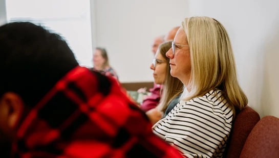 A profile view of a blonde woman in glasses and a striped shirt, sitting in an audience and listening intently. Other people are blurred in the background.