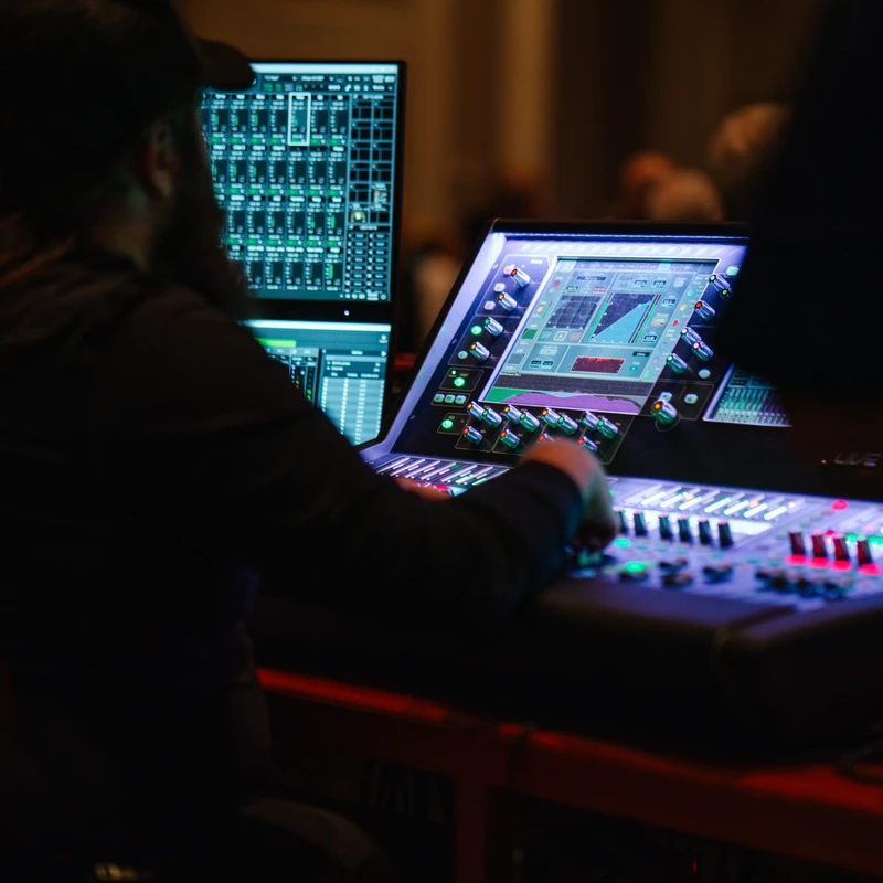 A sound engineer with long hair and a cap, seen from behind, operates a large, glowing audio mixing board and computer screens in a dark room.