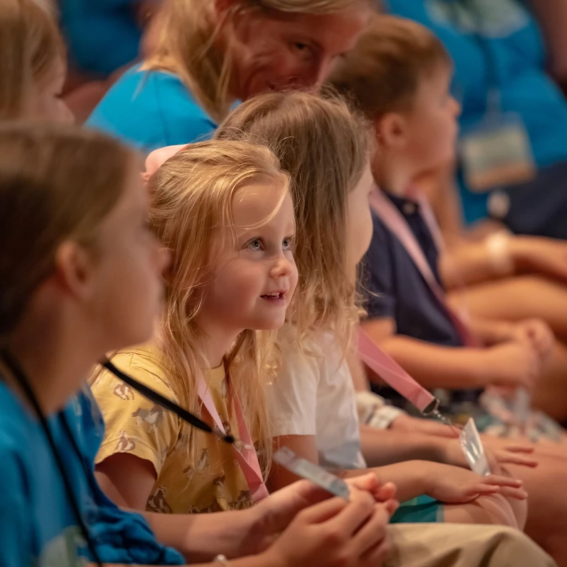 A young blonde girl with a look of wonder on her face sits in an audience, looking towards the right. Other children and adults surround her.