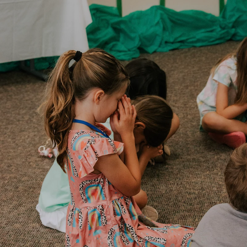 A young girl in a pink rainbow-print dress sits on a carpeted floor, whispering into the ear of another child during a group activity.