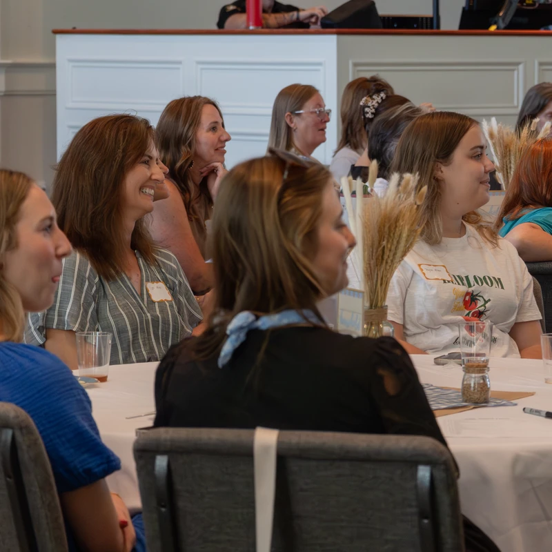 A group of women sit at round tables with white tablecloths, smiling and listening intently during a presentation in a brightly lit conference room.
