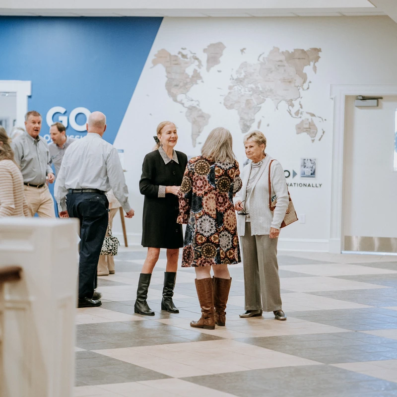 Groups of people stand and socialize in a spacious hall with a checkered floor. A large world map and the word 'GO' are on the wall in the background.