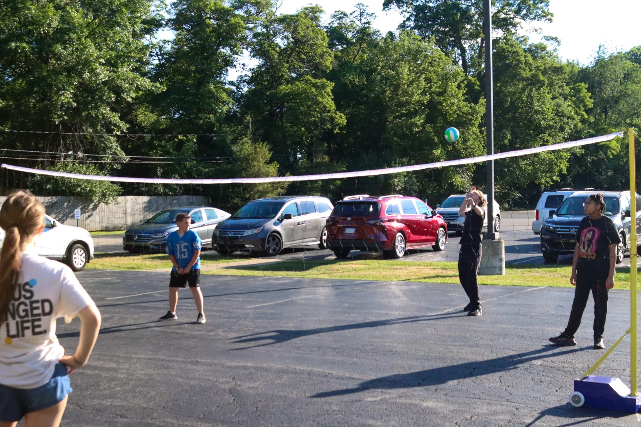 Four young people play volleyball in a paved parking lot on a sunny day. They are using a portable net, with cars and green trees in the background.