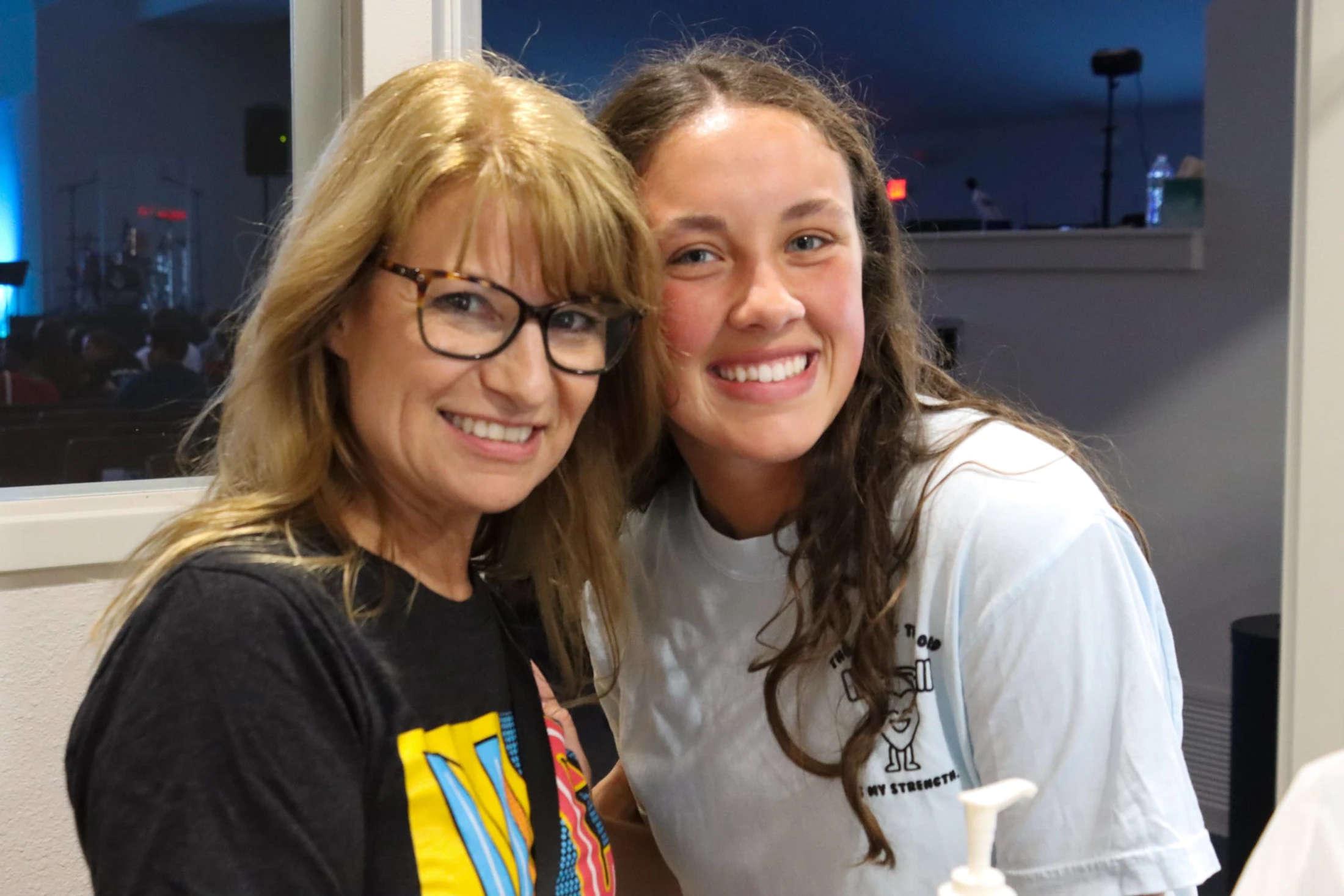 A blonde woman with glasses and a younger woman with long brown hair pose closely together, smiling warmly for the camera in an indoor setting.
