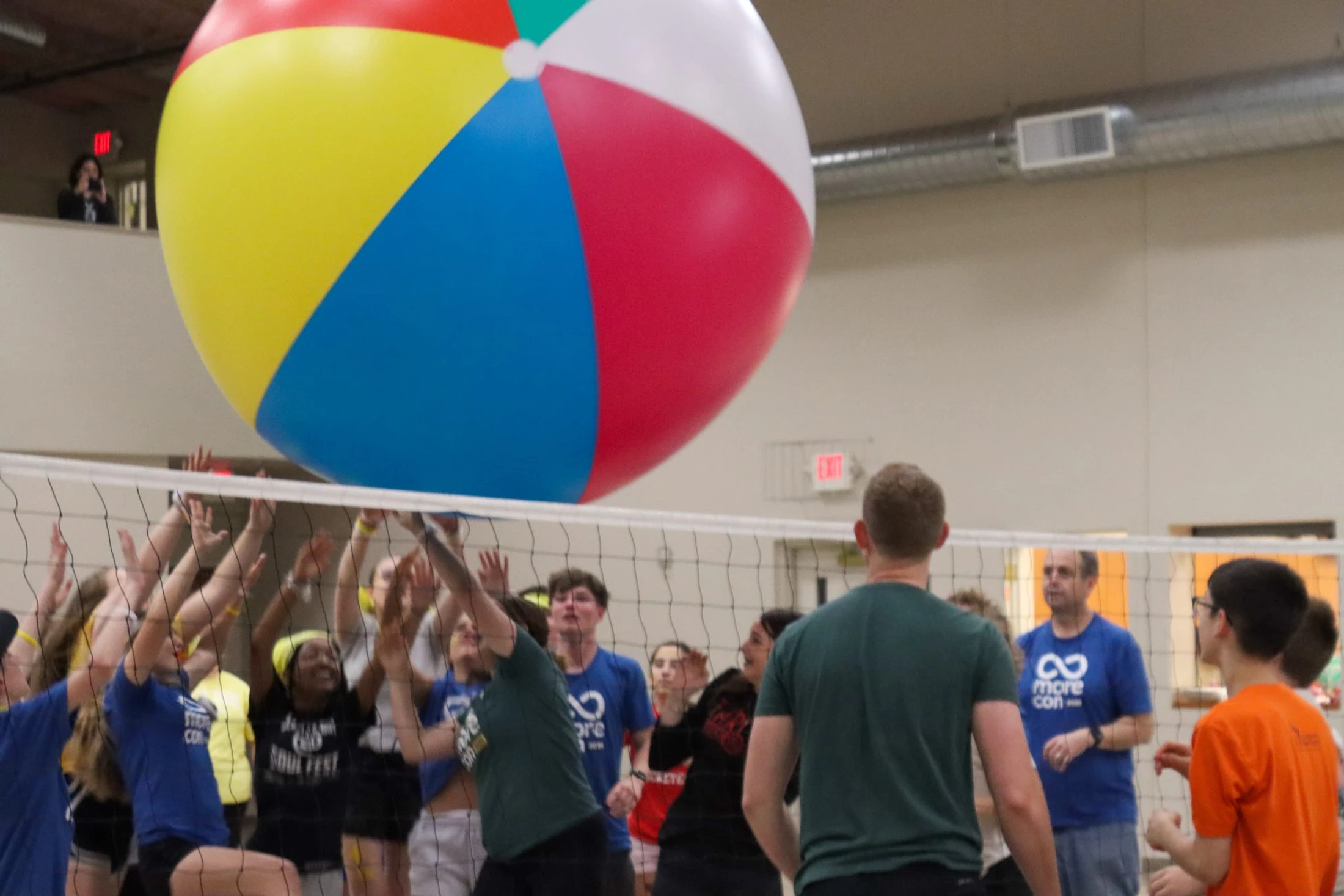 A large group of people play volleyball indoors with a giant, multi-colored beach ball that is floating over the net as they reach up to hit it.