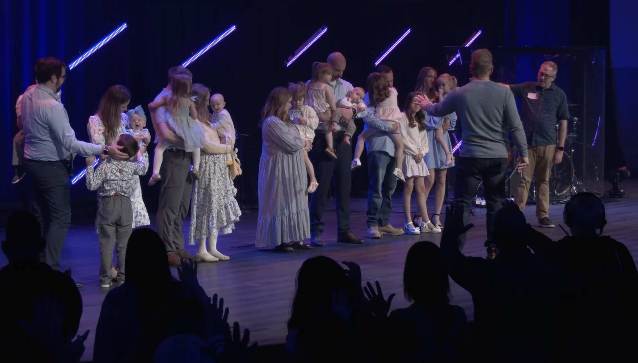 Several families holding babies and children stand on a dimly lit stage as a pastor blesses them, while the audience watches with raised hands.