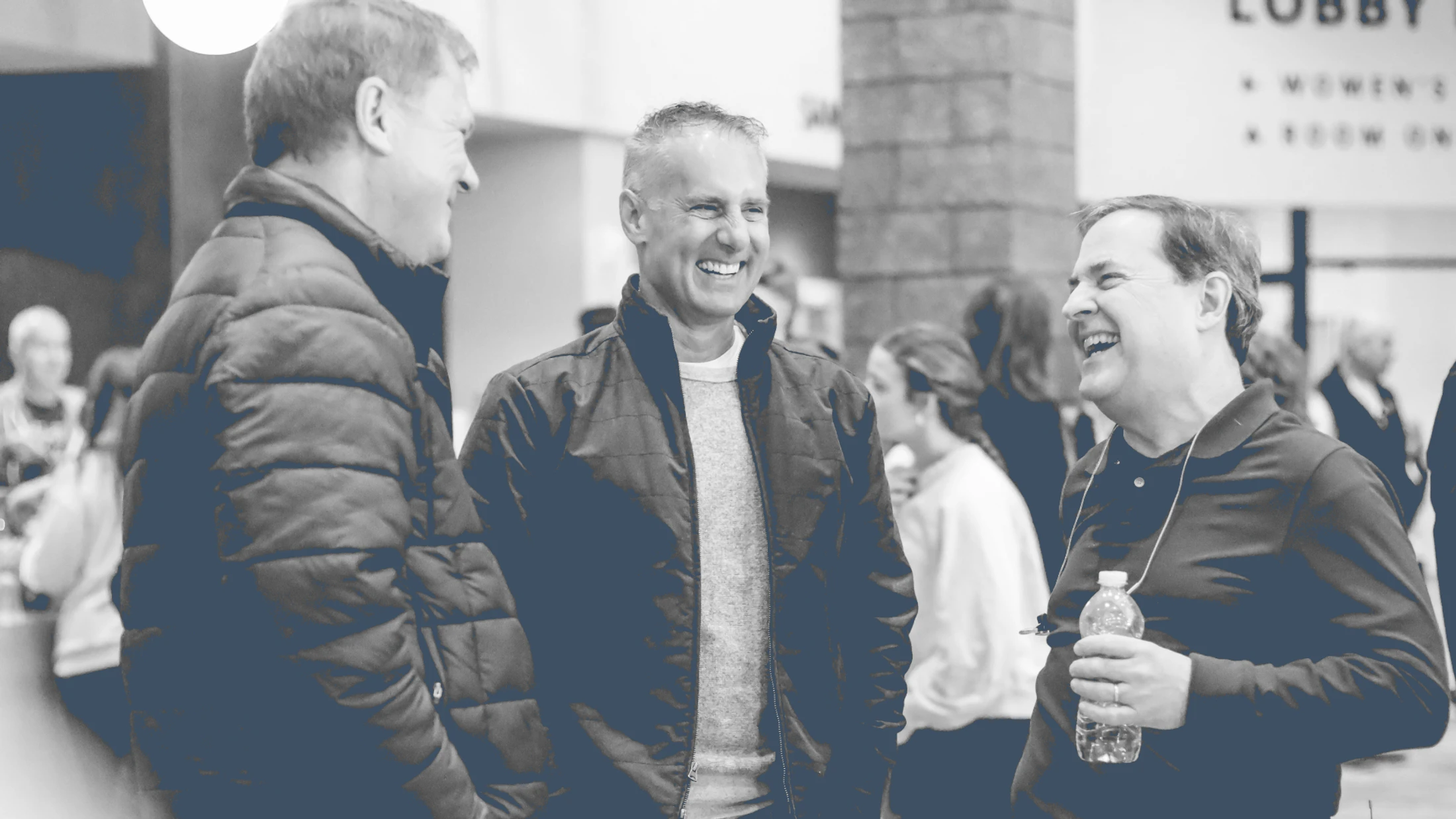 A blue-toned, black and white photo of three middle-aged men standing together, smiling and laughing during a conversation in a busy indoor lobby.