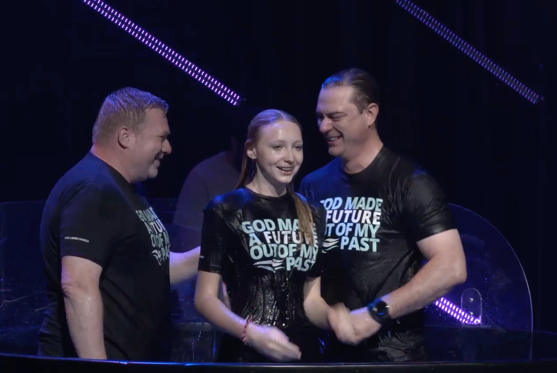 A smiling young woman stands between two smiling men in a baptismal font, all wearing wet black t-shirts after being submerged in the water.