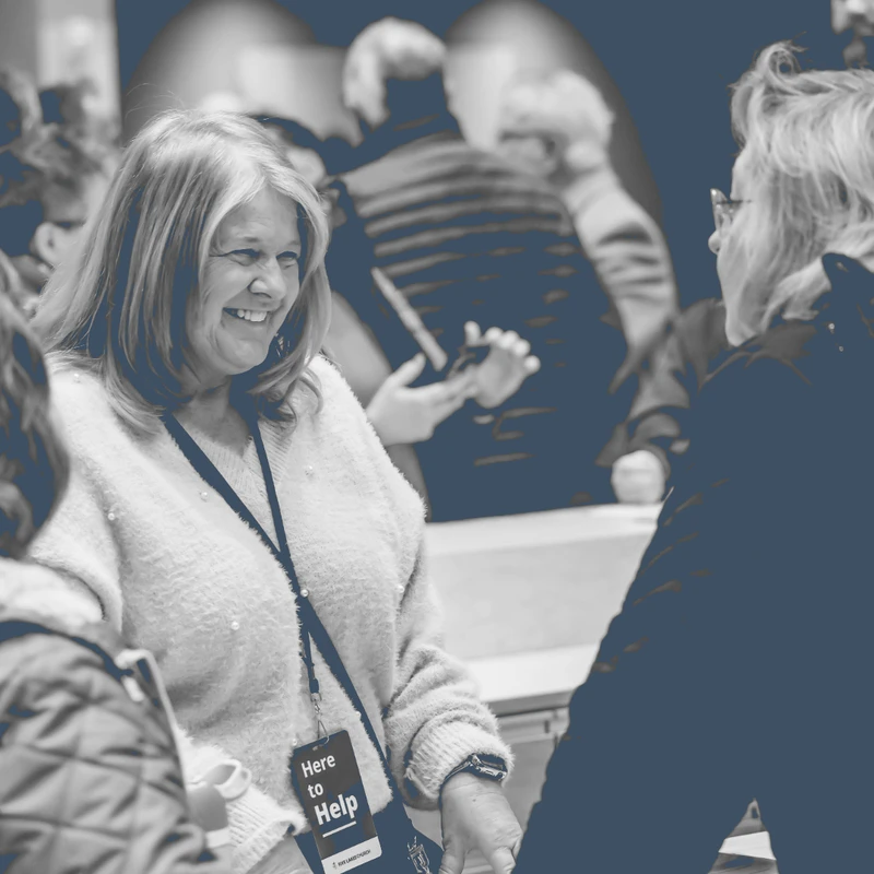 A duotone photo of a smiling woman wearing a lanyard that reads 'Here to Help' while talking with other people in a crowded room.