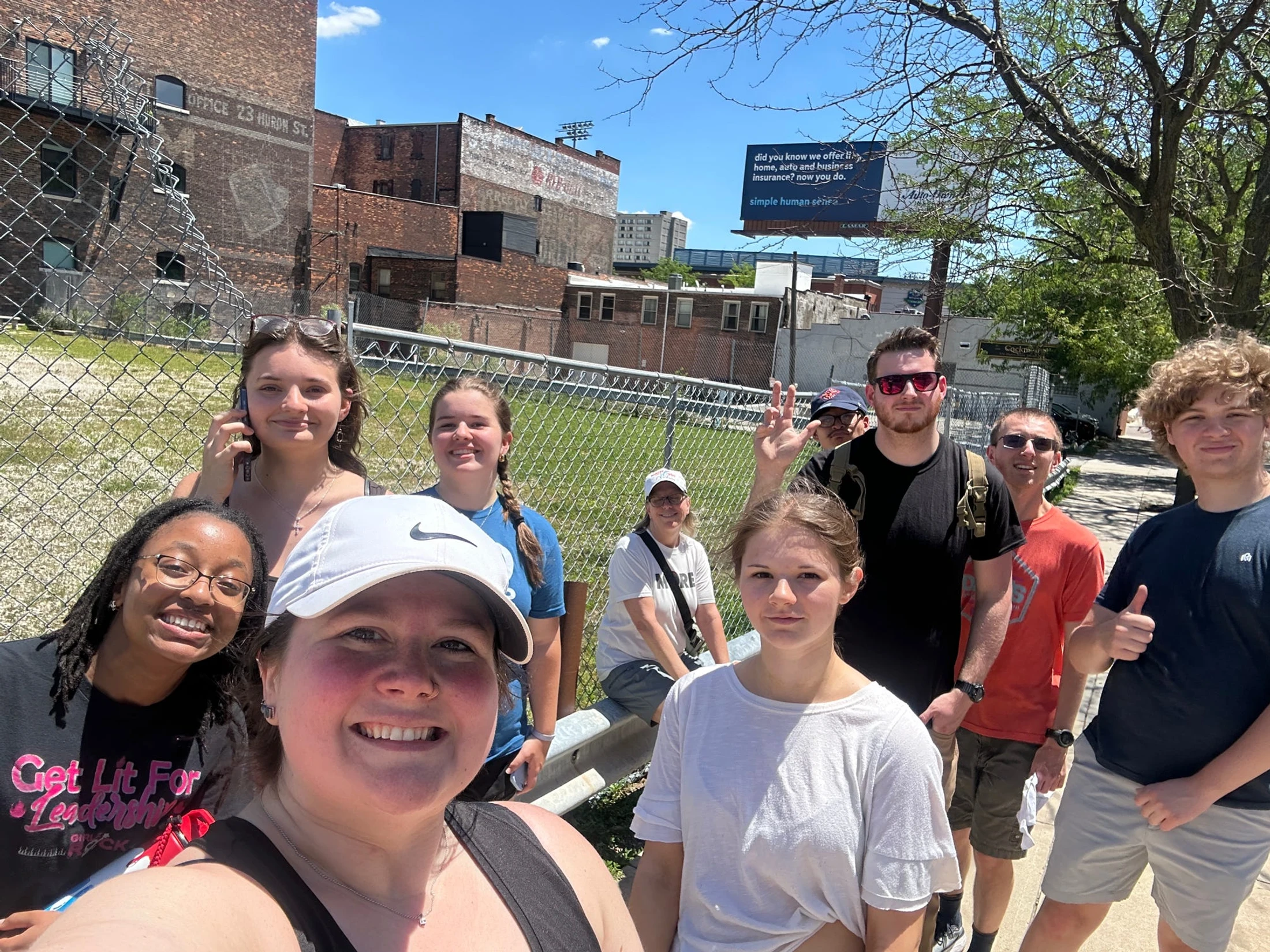A diverse group of about ten people smiling and posing for a selfie outdoors on a sunny day, with a chain-link fence and brick buildings in the background.