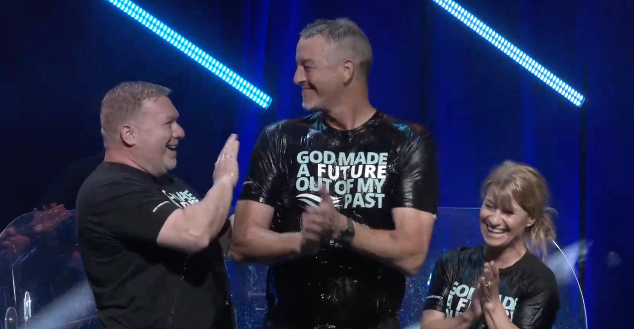 A smiling man, wet from his baptism, wears a t-shirt that reads 'God Made a Future Out of My Past' as two people celebrate with him on a stage.