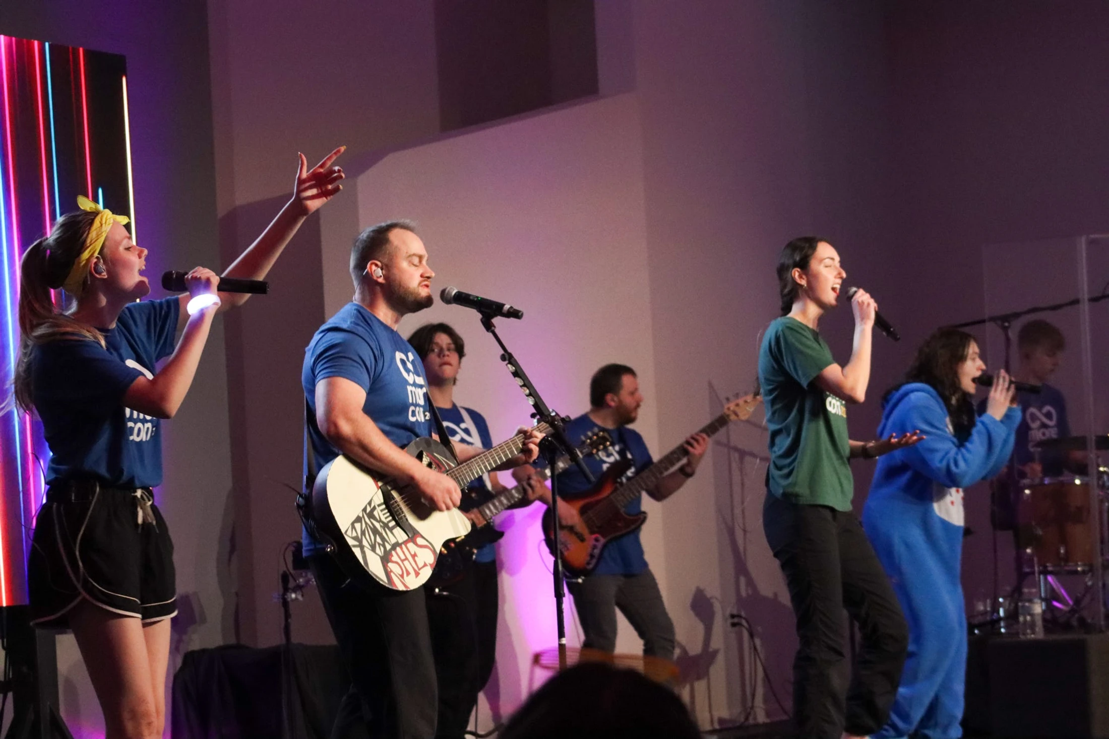 A band with several singers and musicians performs enthusiastically on a stage lit with colorful neon and purple lights.