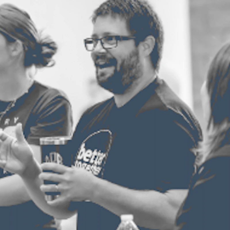 A blue-tinted photo of a man with glasses and a beard smiling and talking to a group. A young woman next to him listens intently.