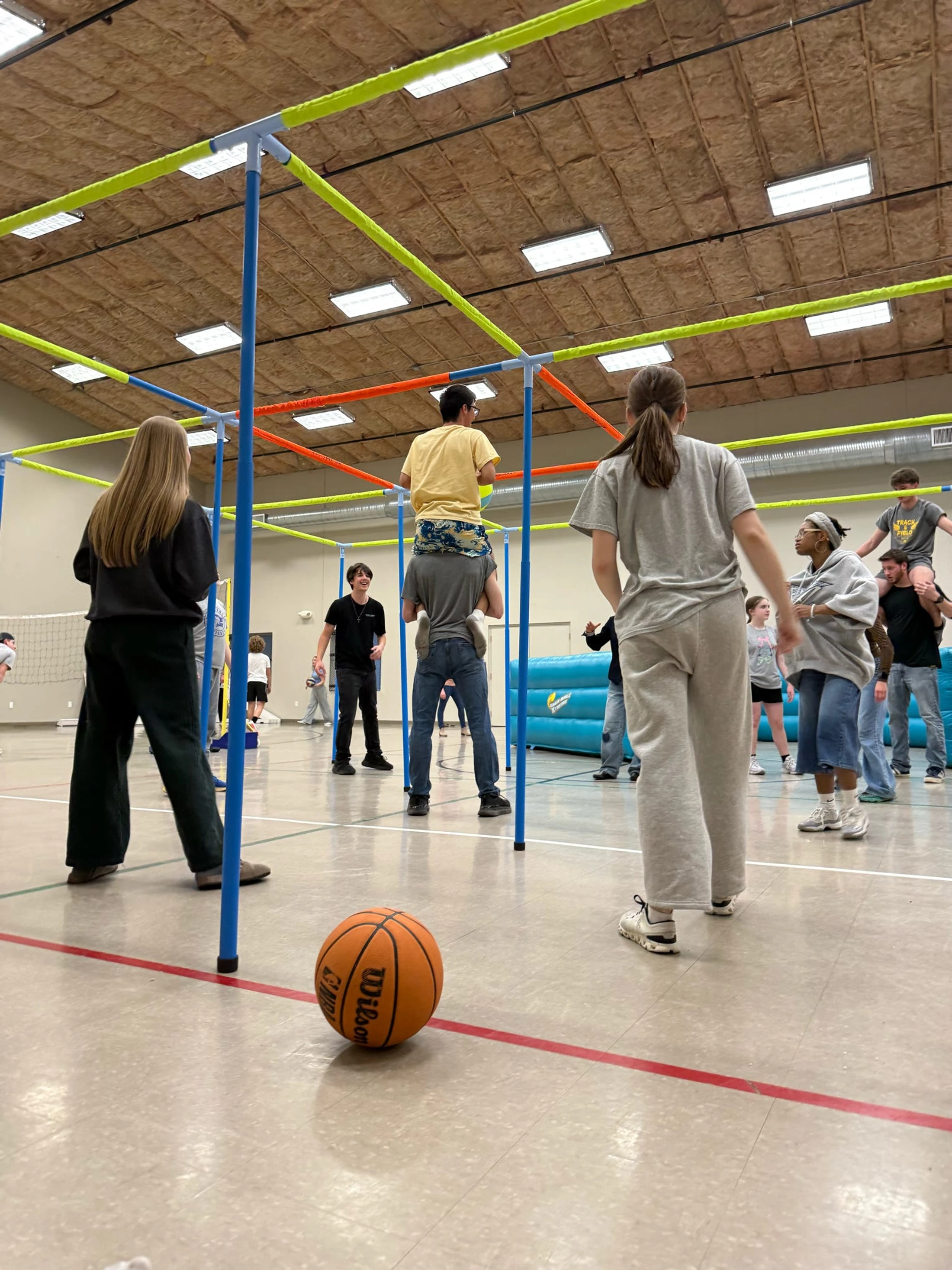 A group of young people in a gymnasium play a game involving a colorful cube-like structure, with some riding on others' shoulders. A basketball sits nearby.