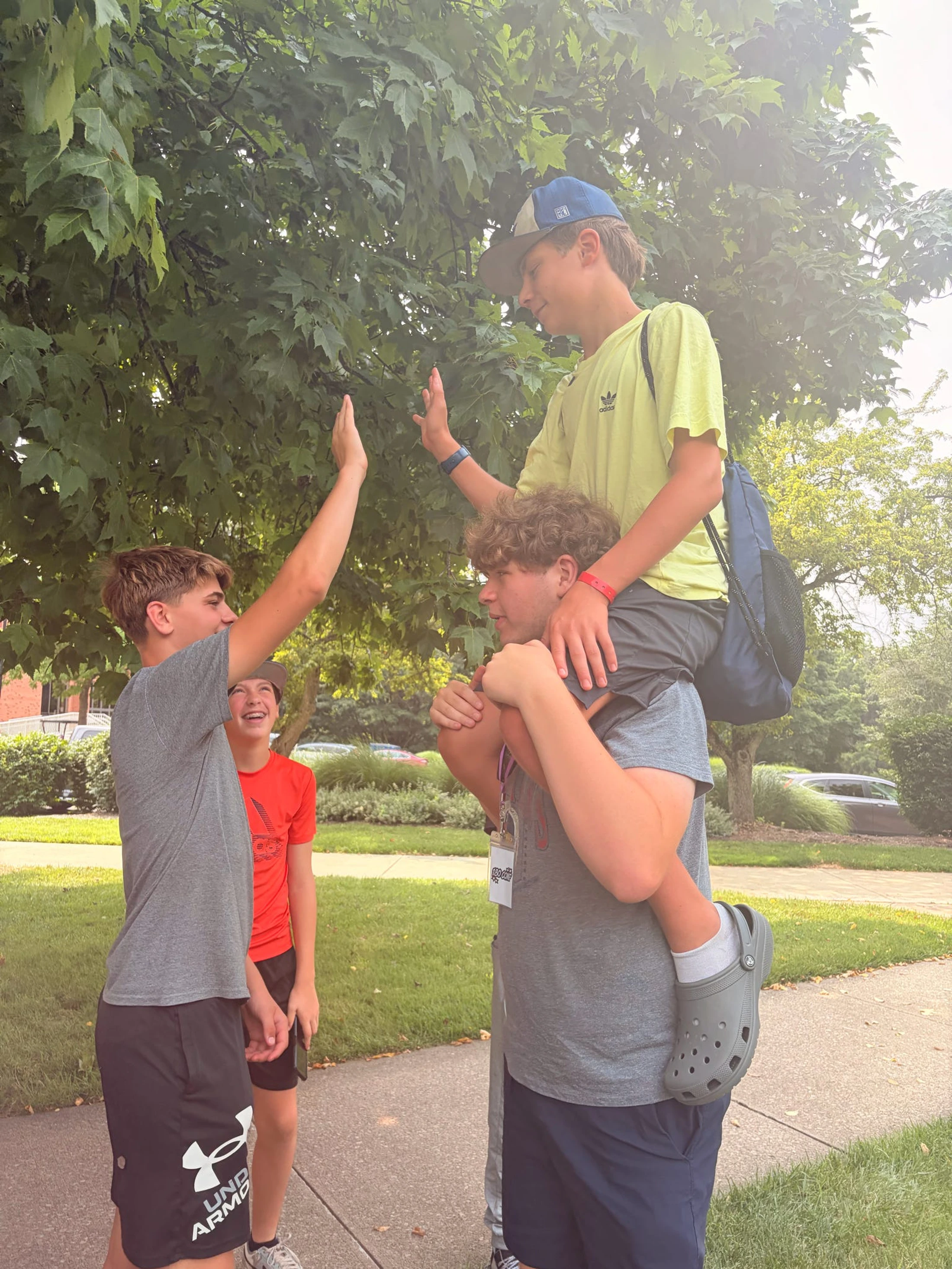 Four teenage boys are outside; one is sitting on another's shoulders and giving a high-five to a third boy, while a fourth boy smiles in the background.