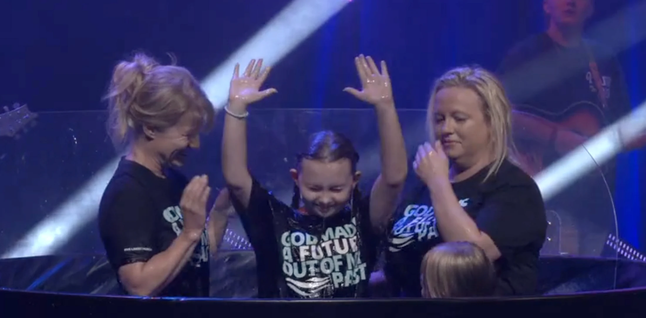 A young girl with wet hair smiles and raises her hands in the air while standing in a baptismal font, flanked by two smiling women on a lit stage.
