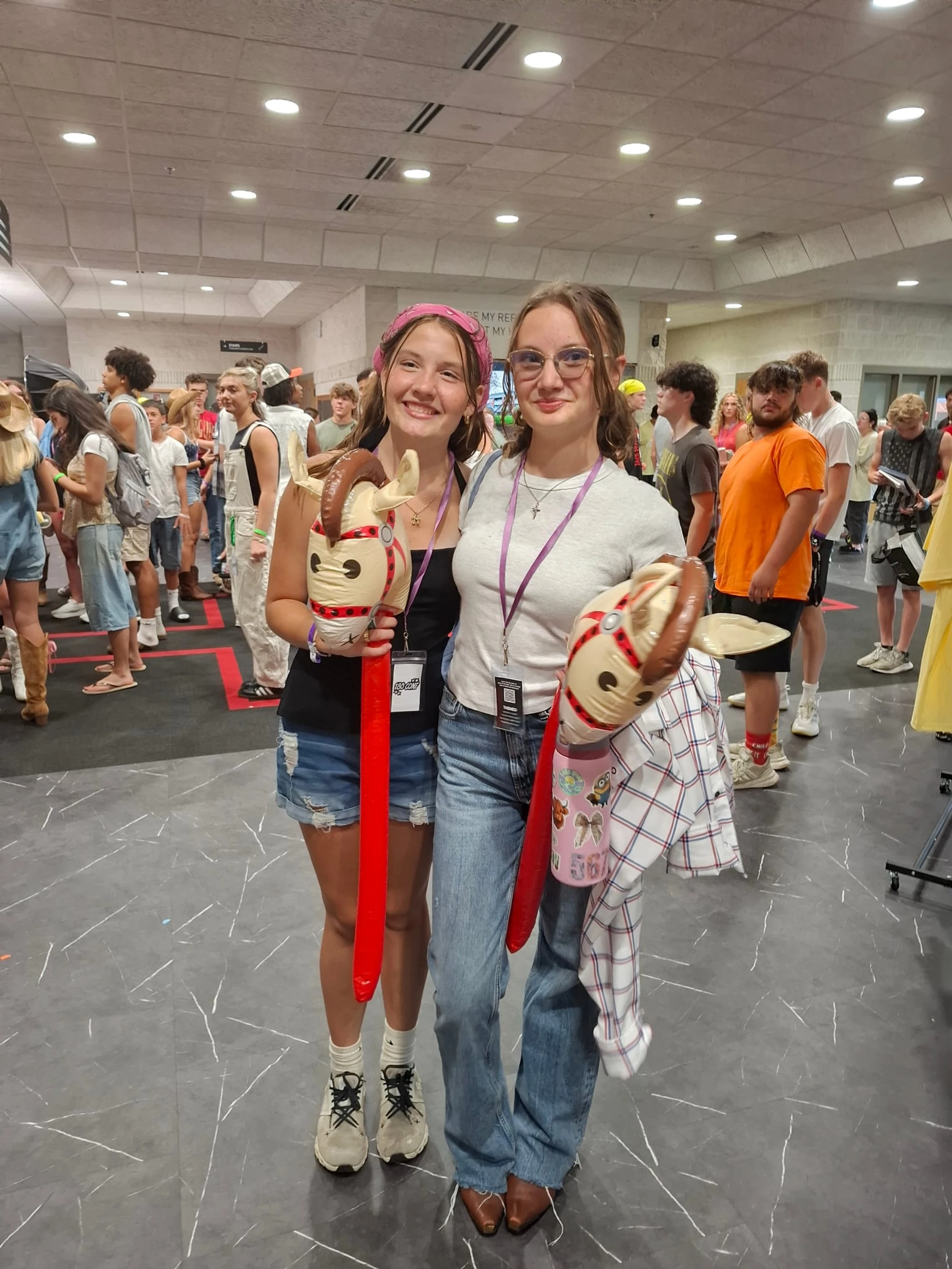 Two smiling young women pose together in a crowded hall, each holding an inflatable toy featuring a goat's head on a long red stick.