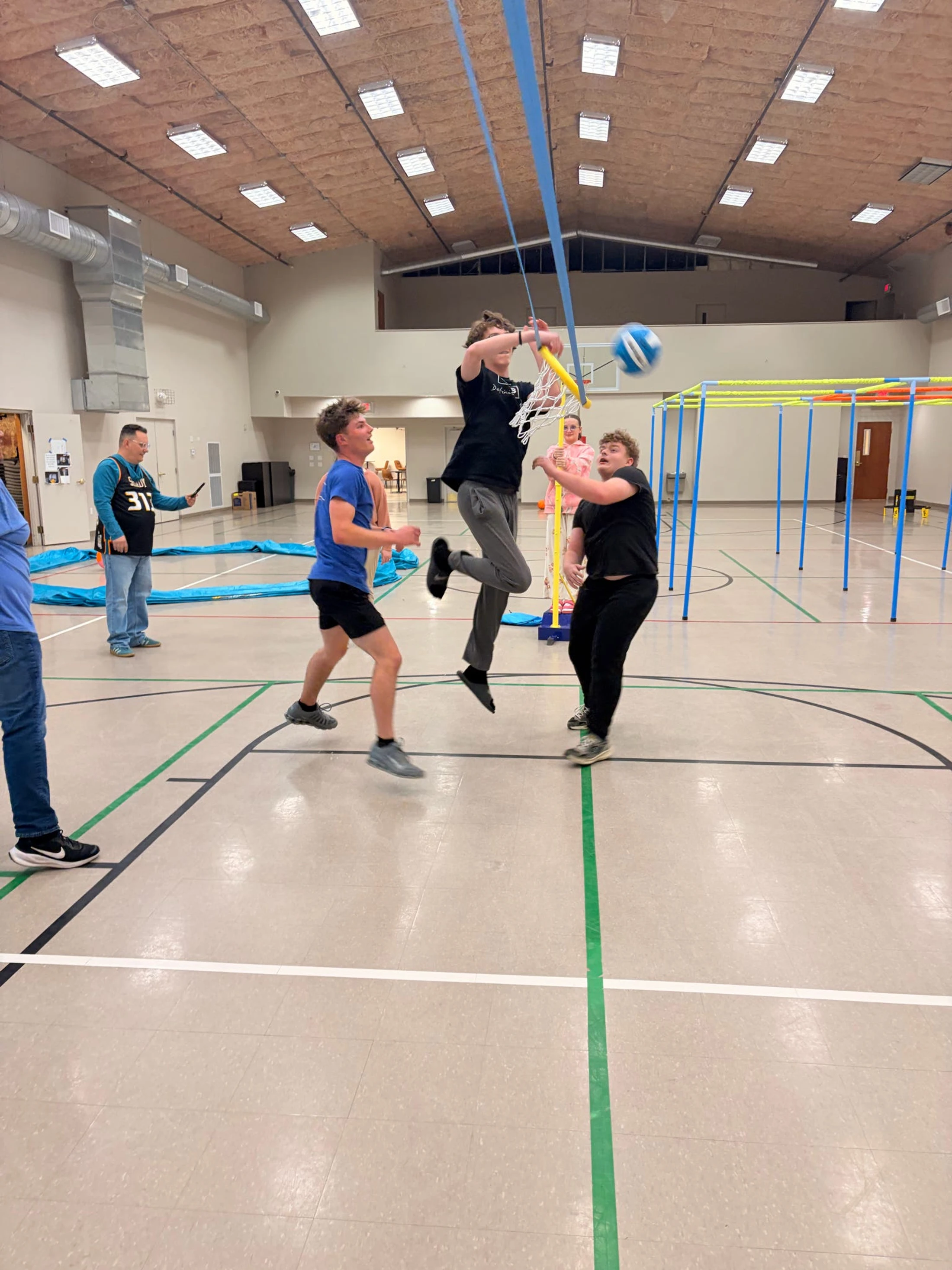 A group of teenagers play an active game on a gym floor. One boy jumps in mid-air to spike a blue and white ball towards a portable yellow hoop.