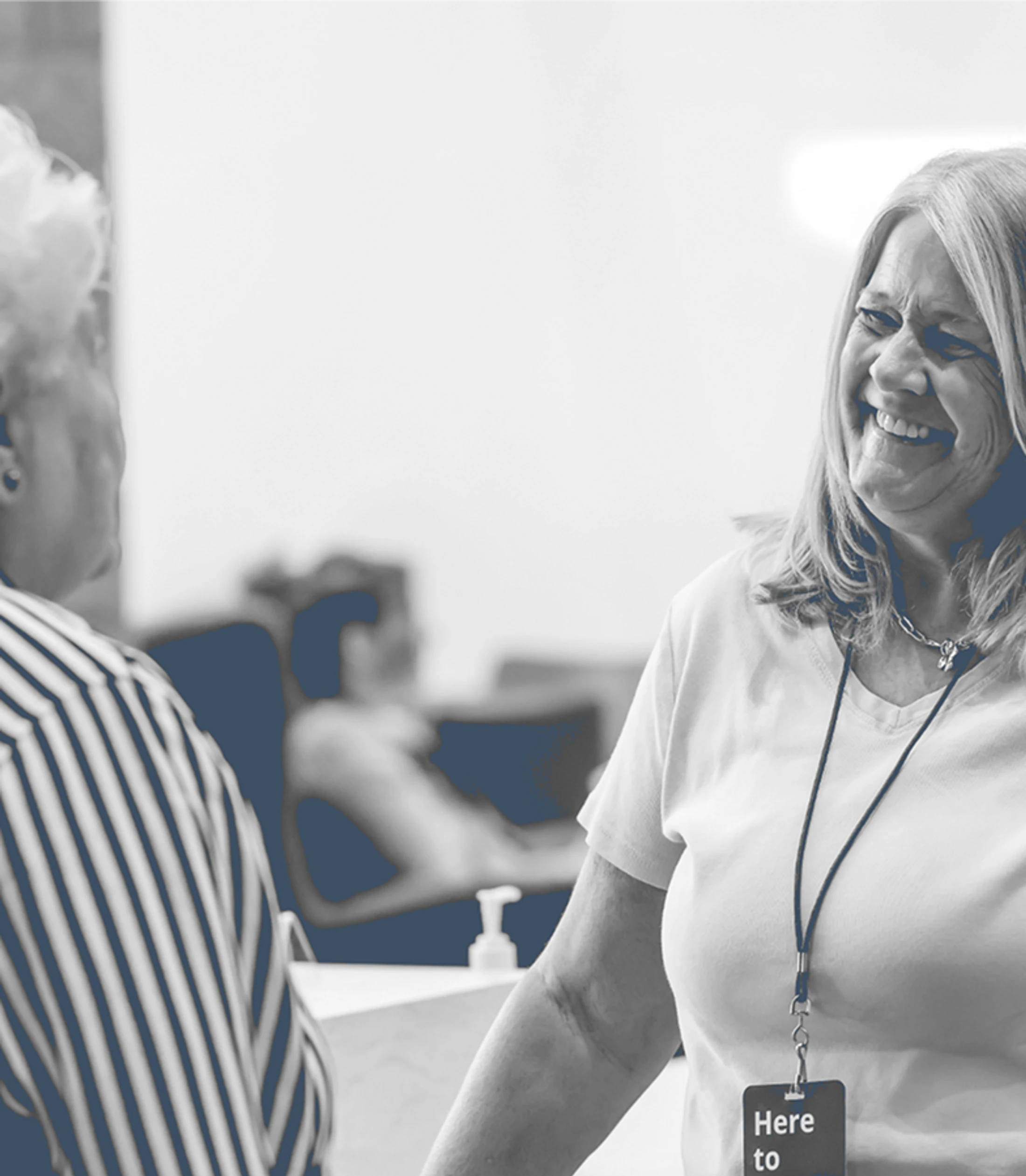 A black and white photo of a smiling woman wearing a lanyard and a badge that says 'Here to', as she speaks to another person at a desk.