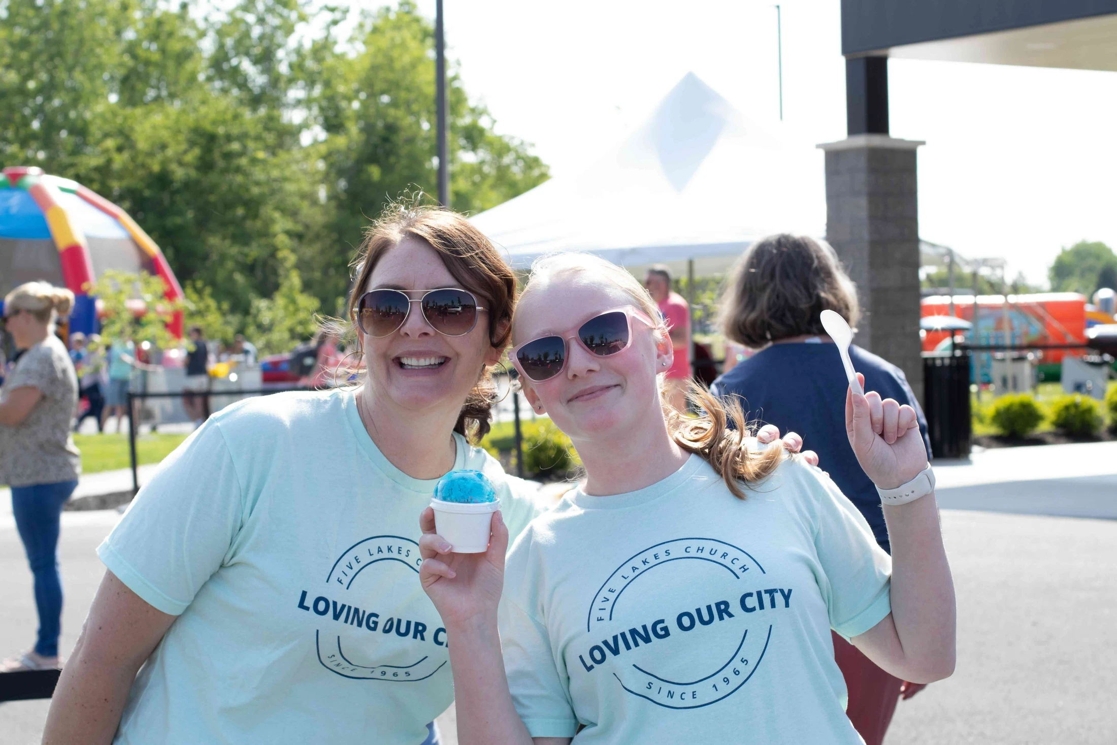 A woman and a girl in matching light blue t-shirts smile at an outdoor event. The girl holds a cup of blue shaved ice and a spoon.