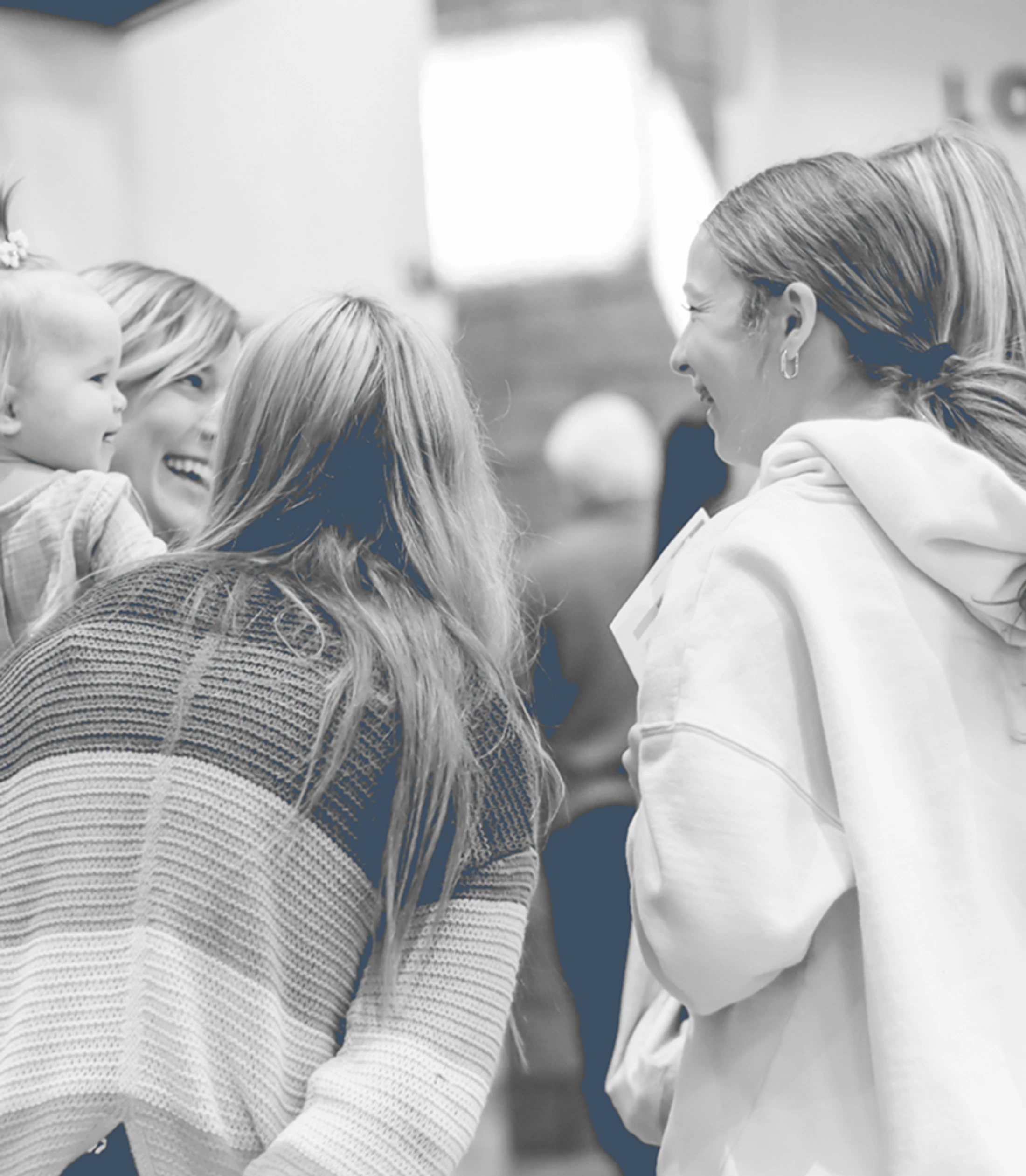 A black and white photo of several women and a baby interacting. A woman on the left holds the baby while smiling at another woman.