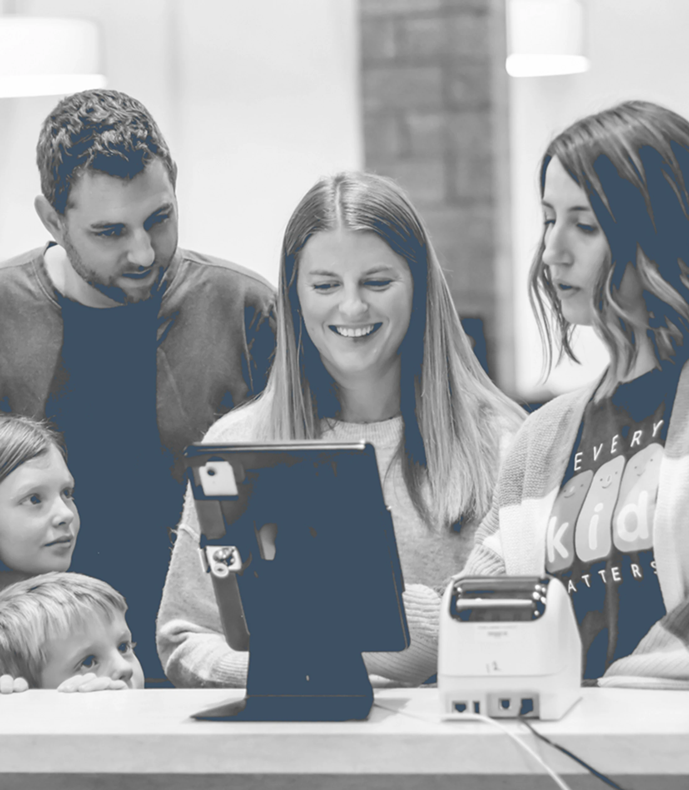 A duotone photo of a family with two children checking in at a welcome desk using a tablet, with a volunteer assisting them.
