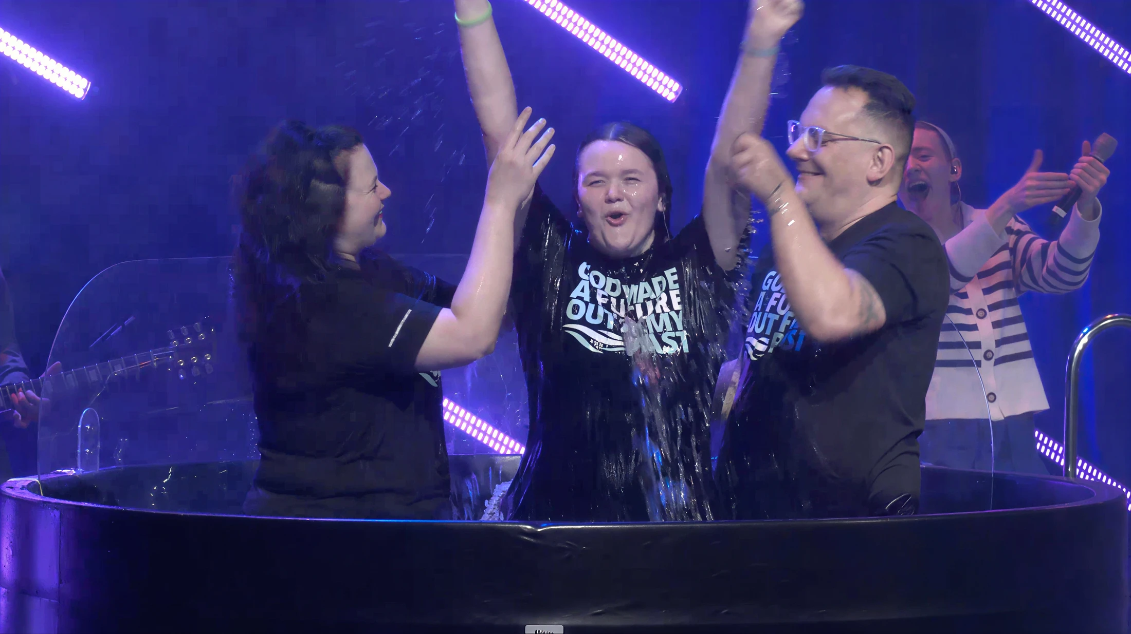 A young person celebrates with arms raised, emerging from a baptismal pool, flanked by a smiling man and woman on a stage with purple lighting.