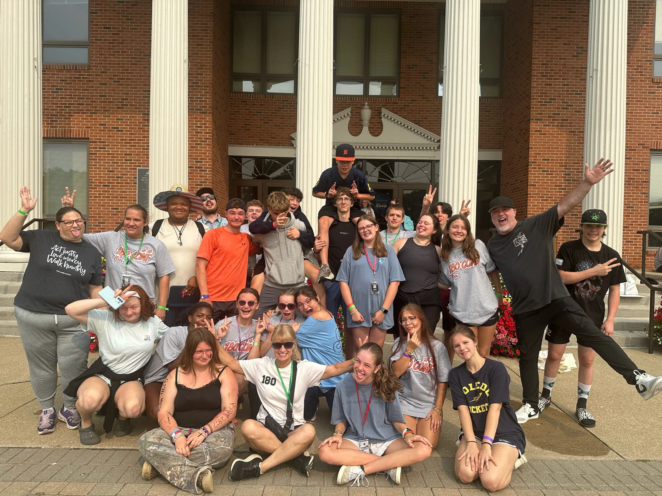 A large group of teenagers and adults pose for a happy, energetic photo on the steps of a brick building with large white columns, many making peace signs.