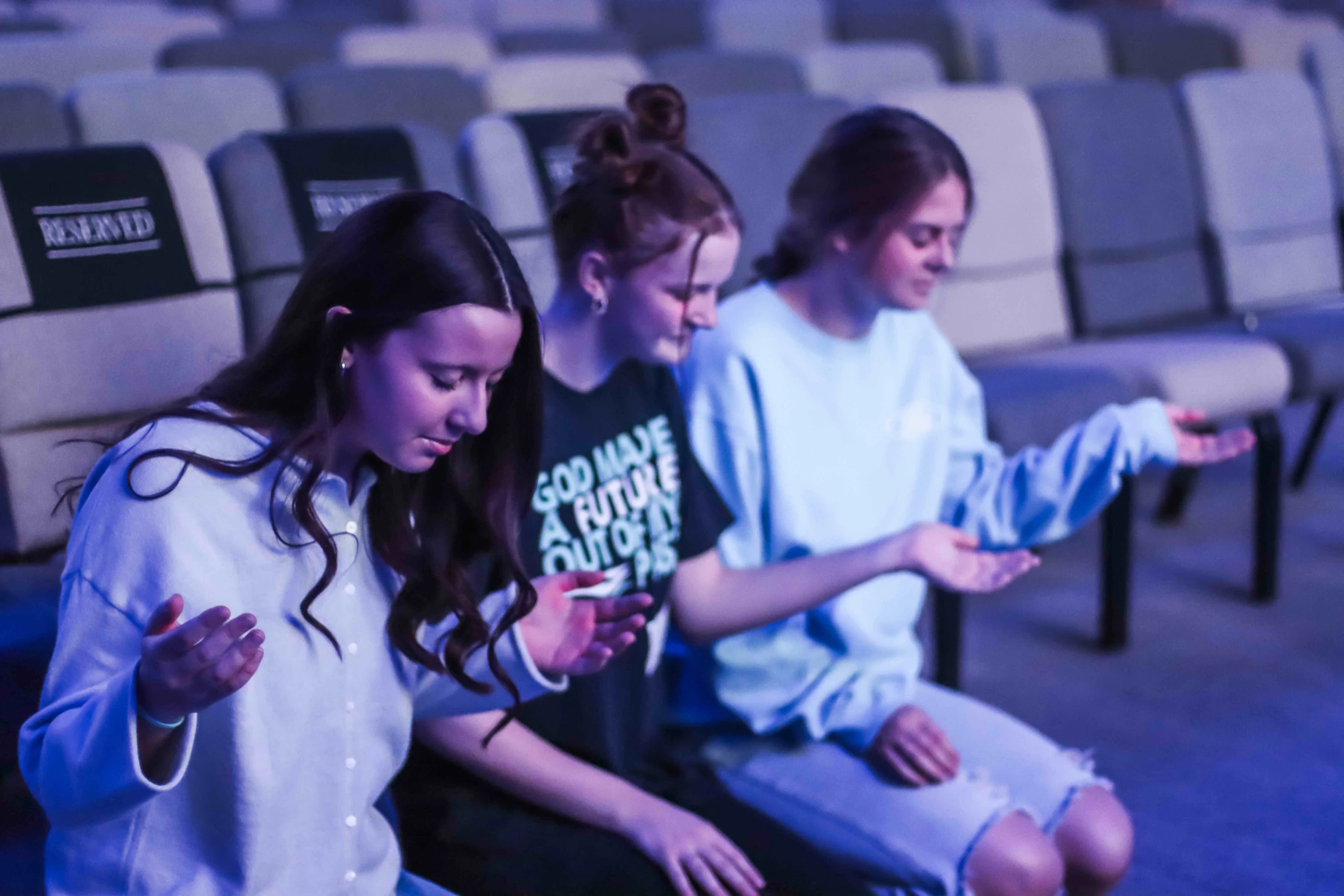 Three young women sit side-by-side in a dimly lit auditorium, their heads bowed and hands open in a gesture of prayer and worship.