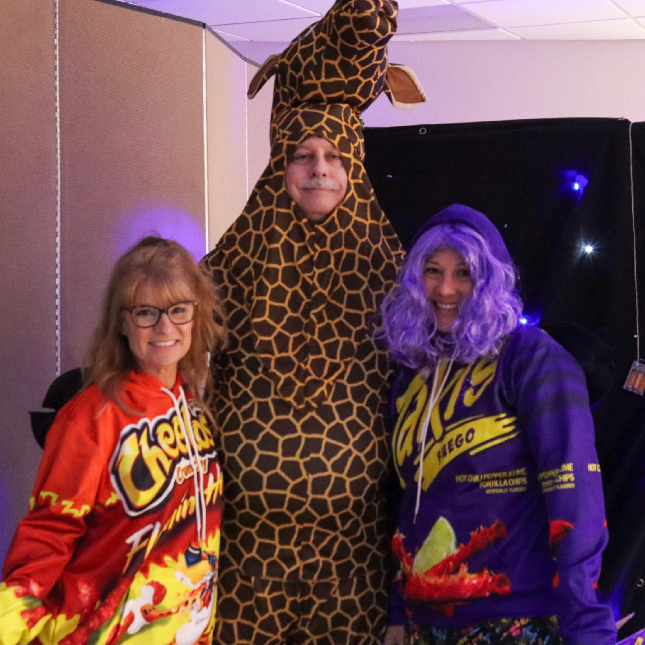 Three adults pose in costumes: a woman in a Cheetos hoodie, a man in a full giraffe suit, and a woman in a Takis hoodie with a bright purple wig.