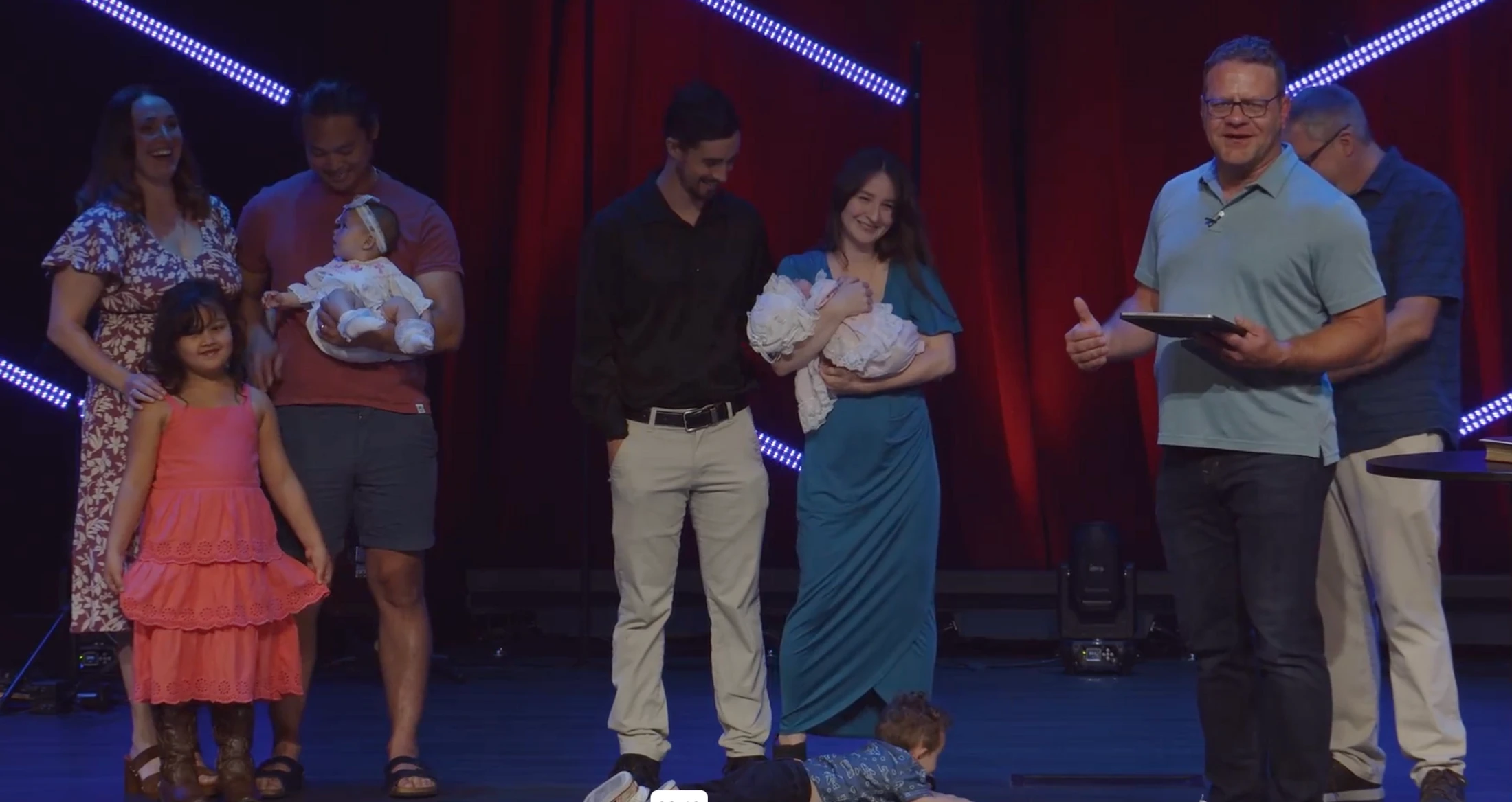 Two families with babies stand on a stage with a pastor during a church ceremony. A young boy lies on the floor while a girl in a pink dress stands nearby.