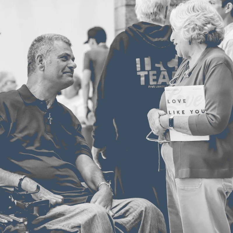A man in a wheelchair smiles while talking to a woman holding a book. The black and white photo has a blue tint, showing a friendly gathering in the background.