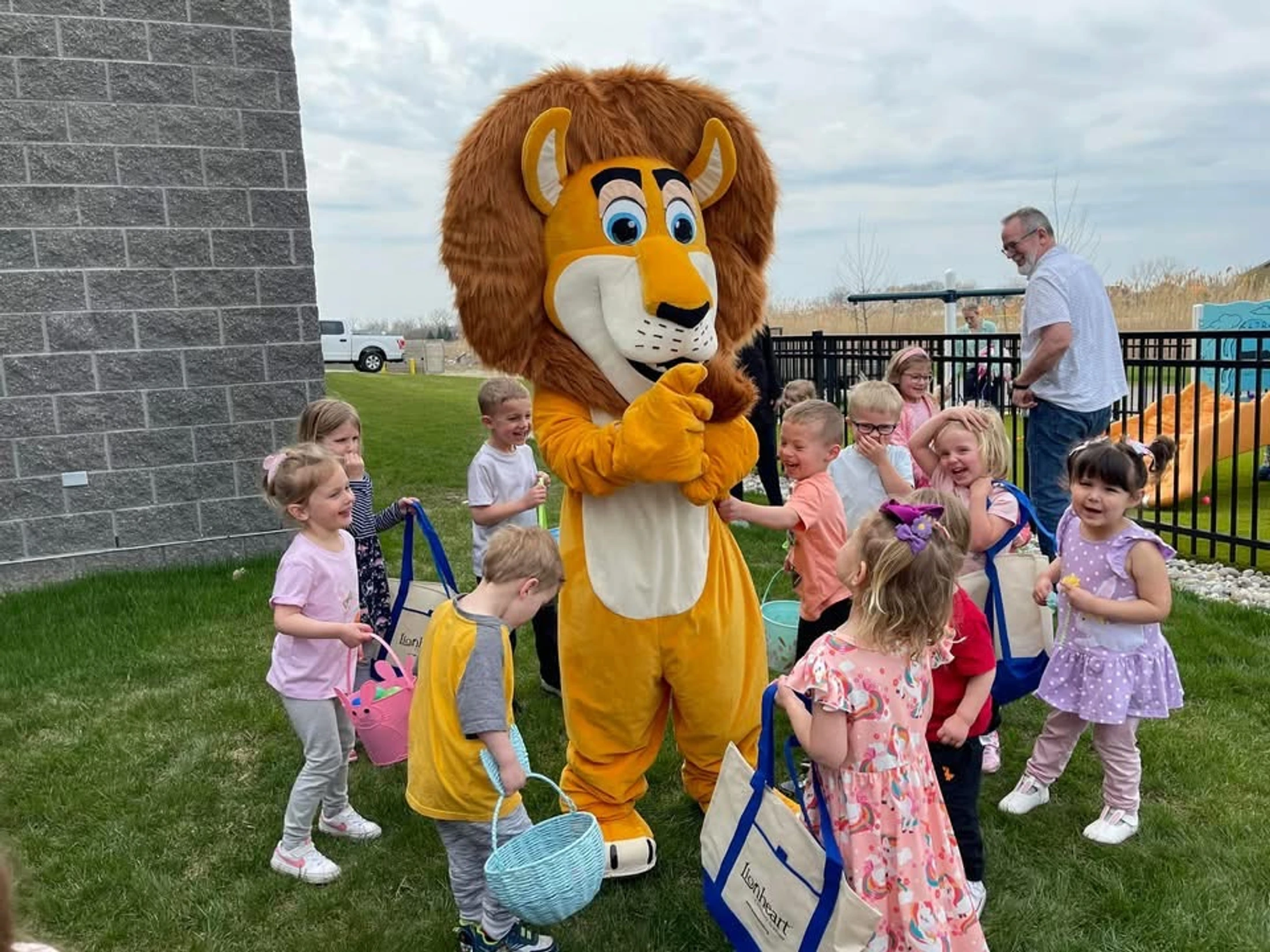 A group of young children with Easter baskets smile as they gather around a person in a friendly lion mascot costume on a grassy lawn.